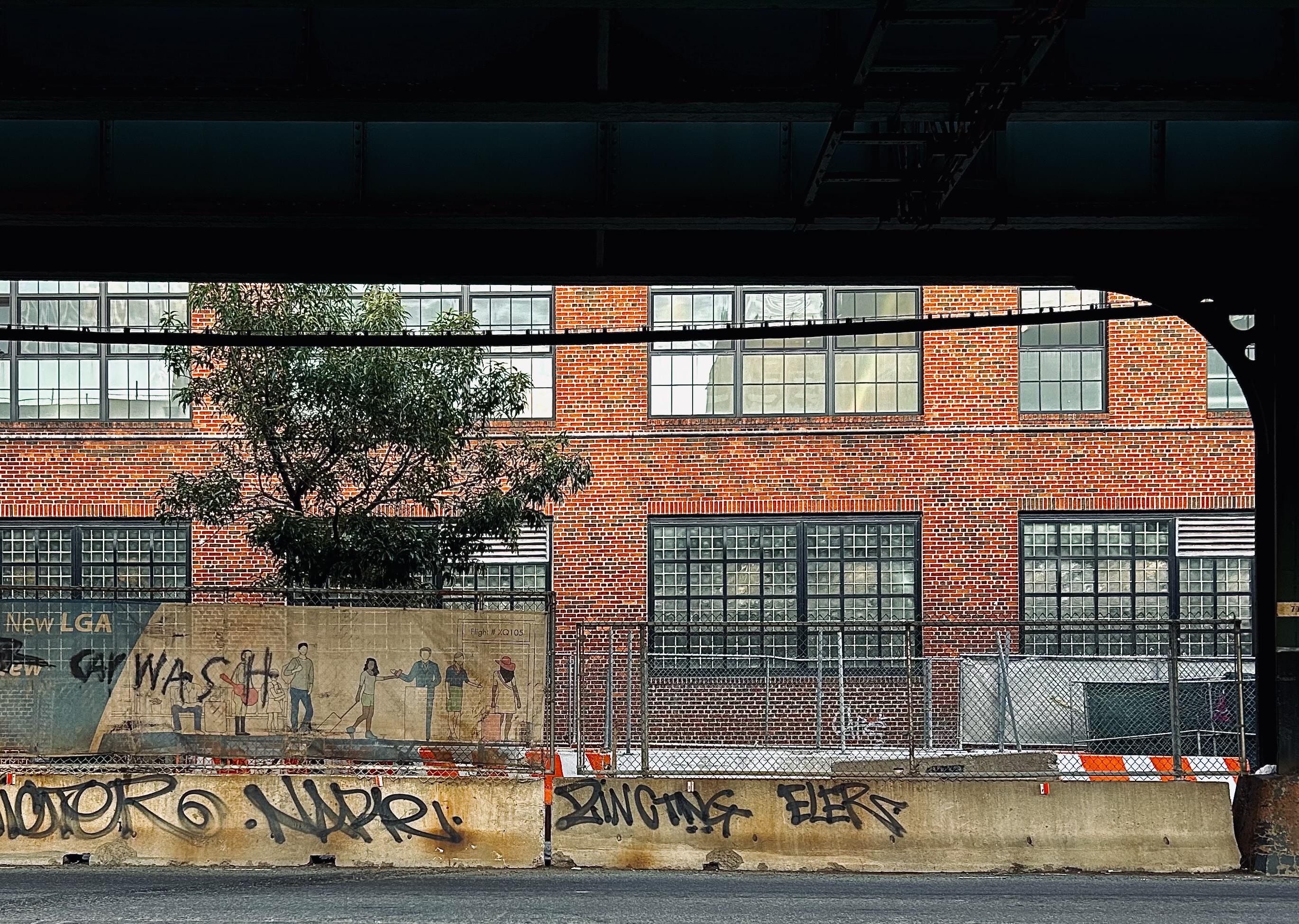 A color, wide-angle photograph that captures the view from beneath the dark, heavy steel girders of an elevated subway track in the Inwood neighborhood of Manhattan. It reveals a sunlit industrial scene. In the background, a large multi-story building made of red and orange bricks features rows of large, multi-paned factory-style windows. A small green tree grows behind a chain-link fence, its leaves partially obscuring the lower windows of the brick facade. The framing of the dark overhead structure creates a heavy, rectangular border at the top of the image, contrasting the shadows of the foreground with the bright textures of the urban backdrop. The mid-ground is dominated by a construction area enclosed by a chain-link fence and tan privacy screening. On the left, the screening features a faded mural of several people walking, adjacent to weathered "CAR WASH" tagging. In front of the fence, a long, weathered concrete barrier runs horizontally across the frame, heavily marked with black spray-painted graffiti tags. Bits of orange and white construction barricades are visible behind the concrete, adding a pop of color to the gritty, utilitarian landscape.