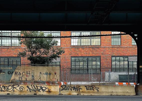 A color, wide-angle photograph that captures the view from beneath the dark, heavy steel girders of an elevated subway track in the Inwood neighborhood of Manhattan. It reveals a sunlit industrial scene. In the background, a large multi-story building made of red and orange bricks features rows of large, multi-paned factory-style windows. A small green tree grows behind a chain-link fence, its leaves partially obscuring the lower windows of the brick facade. The framing of the dark overhead structure creates a heavy, rectangular border at the top of the image, contrasting the shadows of the foreground with the bright textures of the urban backdrop. The mid-ground is dominated by a construction area enclosed by a chain-link fence and tan privacy screening. On the left, the screening features a faded mural of several people walking, adjacent to weathered "CAR WASH" tagging. In front of the fence, a long, weathered concrete barrier runs horizontally across the frame, heavily marked with black spray-painted graffiti tags. Bits of orange and white construction barricades are visible behind the concrete, adding a pop of color to the gritty, utilitarian landscape.