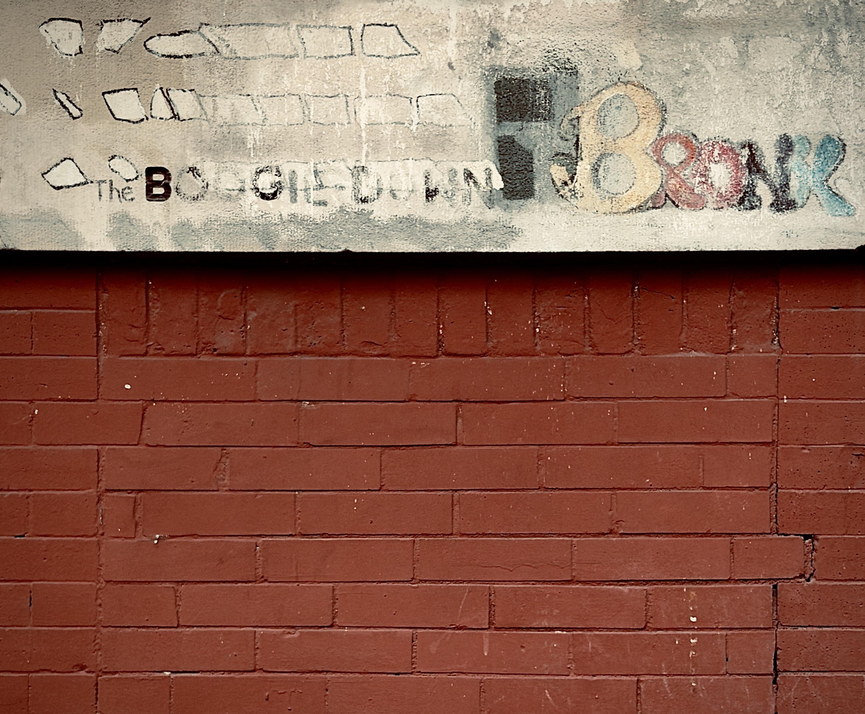 A color, medium-angle photograph in square aspect ratio that features a weathered, horizontal section of a red brick wall topped by a thick, beige-painted concrete ledge. The brickwork is aged with visible cracks and minor wear, while the ledge above displays faded, urban-style lettering and graffiti. On the left side of the ledge, the words "The BOOGIE DOWN" are written in a mix of stenciled and handwritten black text. To the right, the word "BRONX" is rendered in large, stylized bubble letters with a gradient of yellow, red, black, and blue. Above the text, there are faint, sketchy outlines of rectangular shapes resembling windows. The overall aesthetic is gritty and industrial, capturing the textured, layered history of street art in the Bronx.