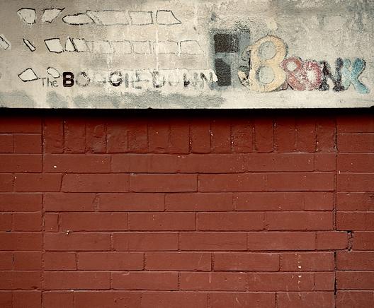 A color, medium-angle photograph in square aspect ratio that features a weathered, horizontal section of a red brick wall topped by a thick, beige-painted concrete ledge. The brickwork is aged with visible cracks and minor wear, while the ledge above displays faded, urban-style lettering and graffiti. On the left side of the ledge, the words "The BOOGIE DOWN" are written in a mix of stenciled and handwritten black text. To the right, the word "BRONX" is rendered in large, stylized bubble letters with a gradient of yellow, red, black, and blue. Above the text, there are faint, sketchy outlines of rectangular shapes resembling windows. The overall aesthetic is gritty and industrial, capturing the textured, layered history of street art in the Bronx.