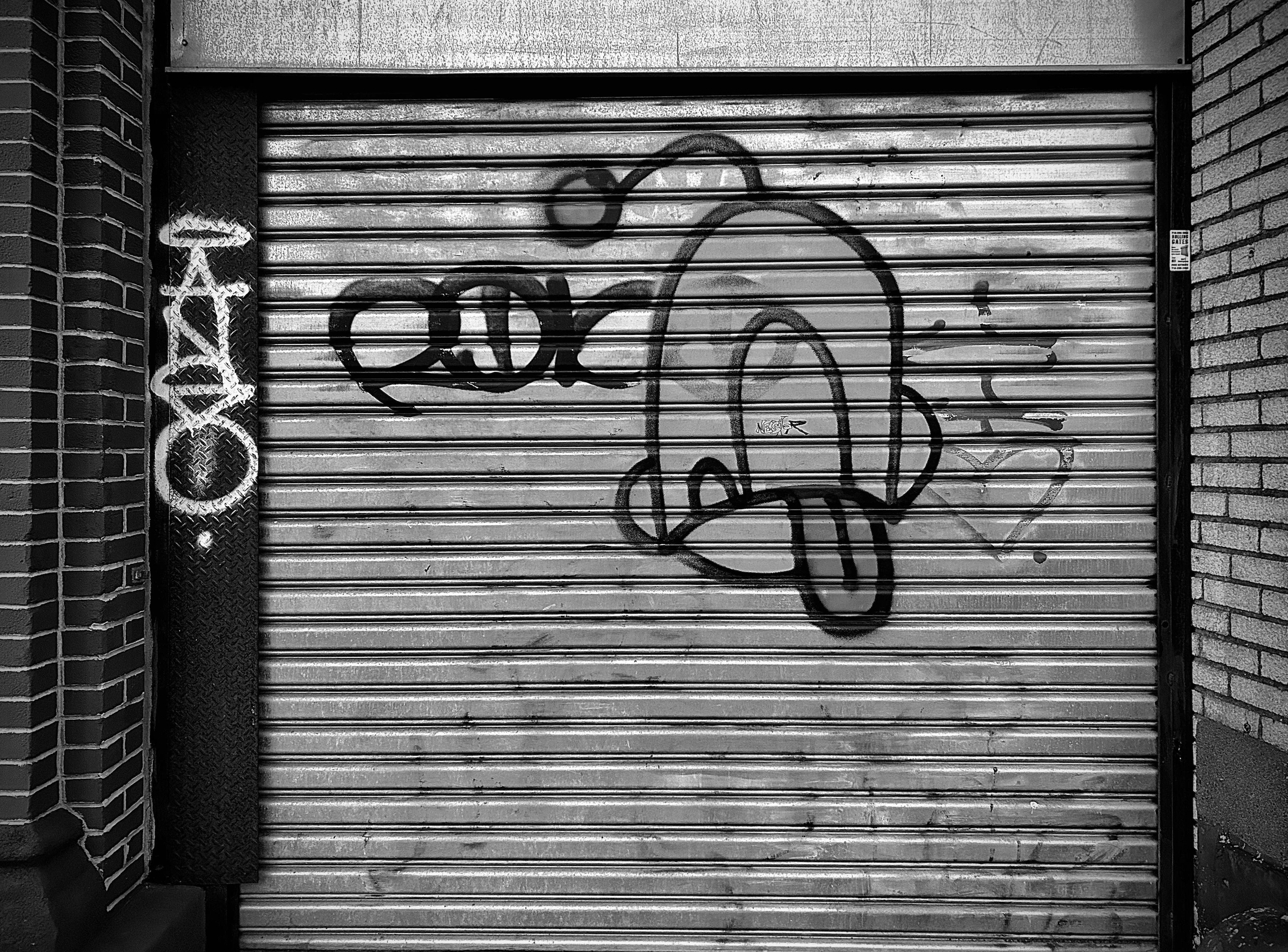 A noir, medium-angle photograph in square aspect ratio that captures a closed metal rolling shutter gate nestled between two brick pillars in downtown Hempstead. The gate is covered in various styles of graffiti, with the most prominent feature being a large, stylized cartoon character rendered in thick, dark spray paint. This character has an oblong, rounded head with a single antenna ending in a circular bulb, and a wide mouth with a protruding tongue, giving it a playful or mischievous expression. To the left of this character is a smaller, horizontal tag consisting of three bold, interlocking letters. On the far left, a vertical strip of diamond-plate metal features a tall, slender tag in white paint that contrasts sharply against the dark background. To the right of the central character, a more delicate, etched-style tag includes a faded small heart at the bottom. The lighting creates a high-contrast effect, emphasizing the horizontal ridges of the shutter and the gritty, urban texture of the surrounding brickwork. The overall composition is centered and symmetrical, focusing entirely on the layered street art of the storefront.