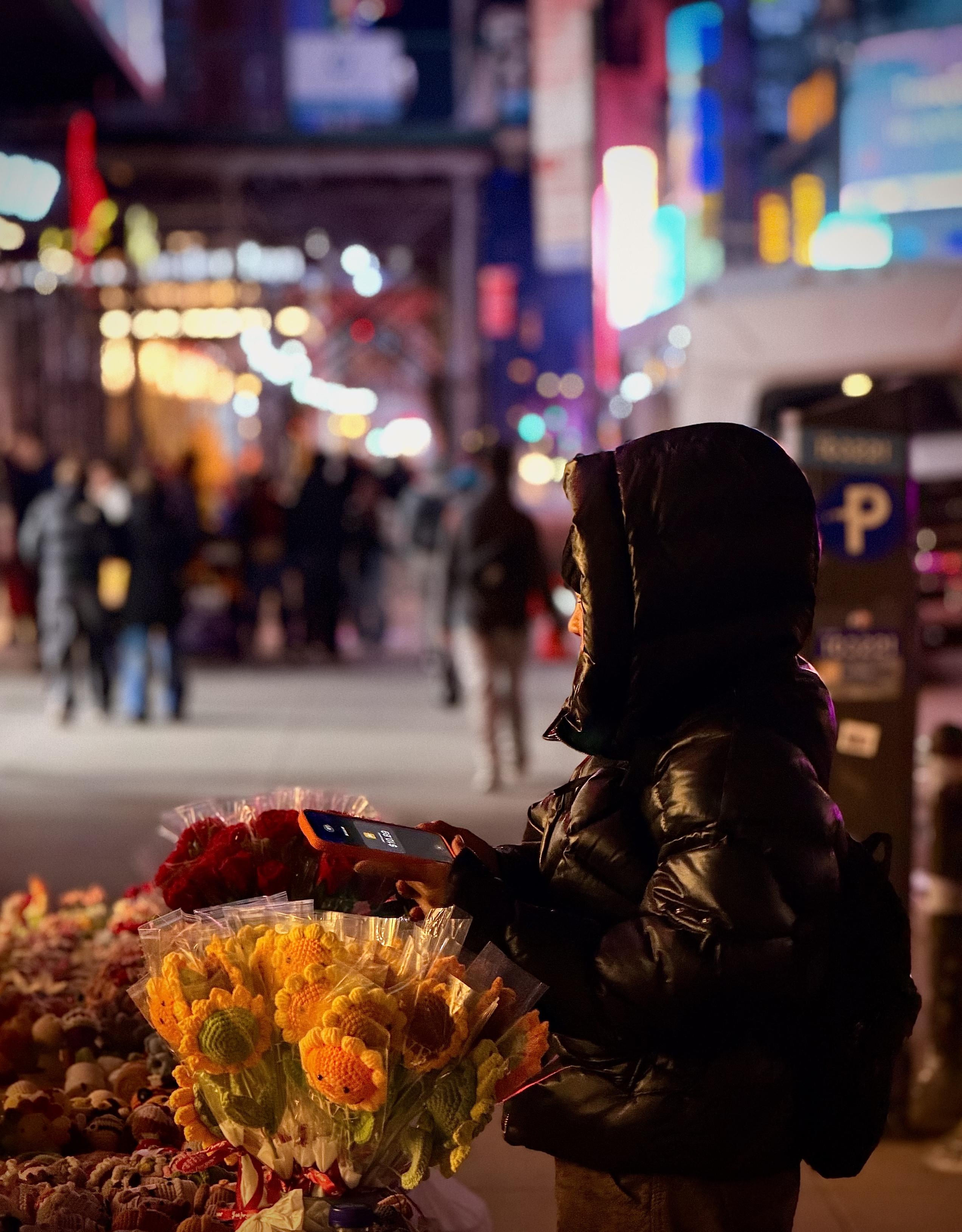 A color, wide-angle portrait style photograph that captures a candid night scene in Times Square, characterized by a vibrant, bokeh-blurred background of neon lights and city activity. In the foreground, a person bundled in a shiny, black puffer jacket with the hood up stands behind a street vendor display. They are holding an orange iPhone 17 Pro horizontally over a colorful arrangement of handcrafted goods, waiting for a contactless payment. The focus is sharp on the vendor and their immediate wares, creating an intimate feel amidst the surrounding metropolitan chaos. The vendor’s table is filled with charming, crocheted fabric flowers wrapped in clear plastic sleeves. Bright yellow sunflowers with smiling orange centers dominate the lower left, while deep red roses are visible just behind them. To the far left, a variety of smaller knitted trinkets and characters are densely packed. On the right, a dark parking meter with a prominent blue "P" sign stands near the vendor, grounding the scene in a public sidewalk setting. The warm glow from the phone screen and nearby streetlights reflects off the vendor's jacket, emphasizing the texture of the fabric and the modern nature of this street-side transaction. The sidewalk is filled without out of focus people standing and walking.