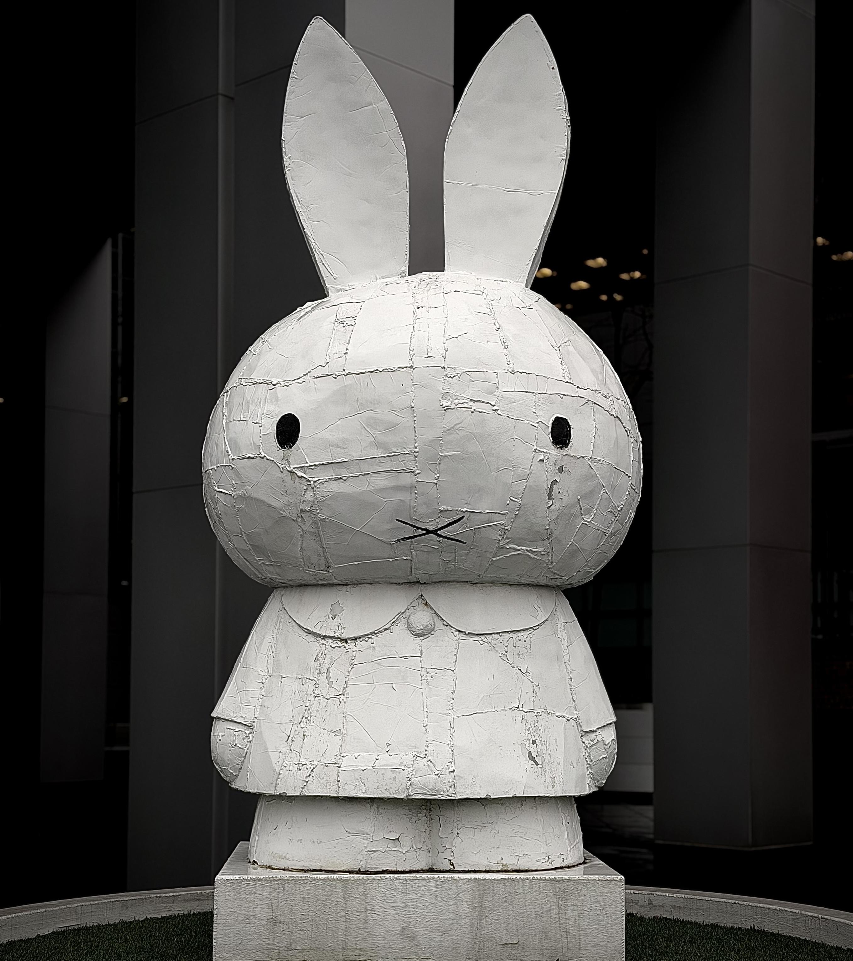 A color, medium-angle photograph in portrait orientation that capture a large-scale, monochromatic white sculpture of the character Miffy, created by artist Tom Sachs installed in lower Manhattan. Standing on a low, square concrete plinth, the sculpture is framed by the dark, towering vertical columns of a modern skyscraper in the background. The statue is made in a bricolage style, where the surface appears intentionally rugged and industrial, covered in a patchwork of textured layers that look like tape, resin, and fiberglass. The rabbit figure has a large rounded head, tall pointed ears, and a simple "X" for a mouth, with two small, dark circular eyes that provide a sharp contrast to the stark white body. The scene is captured with a high-contrast, dramatic aesthetic that emphasizes the gritty texture of the sculpture against the clean, dark lines of the surrounding architecture. A small patch of green artificial turf is visible at the very bottom of the frame, encircling the base of the statue. The lighting is focused, casting subtle shadows within the cracks and seams of the sculpture's surface, which gives the piece a weathered feel.