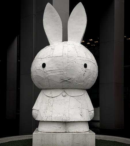 A color, medium-angle photograph in portrait orientation that capture a large-scale, monochromatic white sculpture of the character Miffy, created by artist Tom Sachs installed in lower Manhattan. Standing on a low, square concrete plinth, the sculpture is framed by the dark, towering vertical columns of a modern skyscraper in the background. The statue is made in a bricolage style, where the surface appears intentionally rugged and industrial, covered in a patchwork of textured layers that look like tape, resin, and fiberglass. The rabbit figure has a large rounded head, tall pointed ears, and a simple "X" for a mouth, with two small, dark circular eyes that provide a sharp contrast to the stark white body. The scene is captured with a high-contrast, dramatic aesthetic that emphasizes the gritty texture of the sculpture against the clean, dark lines of the surrounding architecture. A small patch of green artificial turf is visible at the very bottom of the frame, encircling the base of the statue. The lighting is focused, casting subtle shadows within the cracks and seams of the sculpture's surface, which gives the piece a weathered feel.