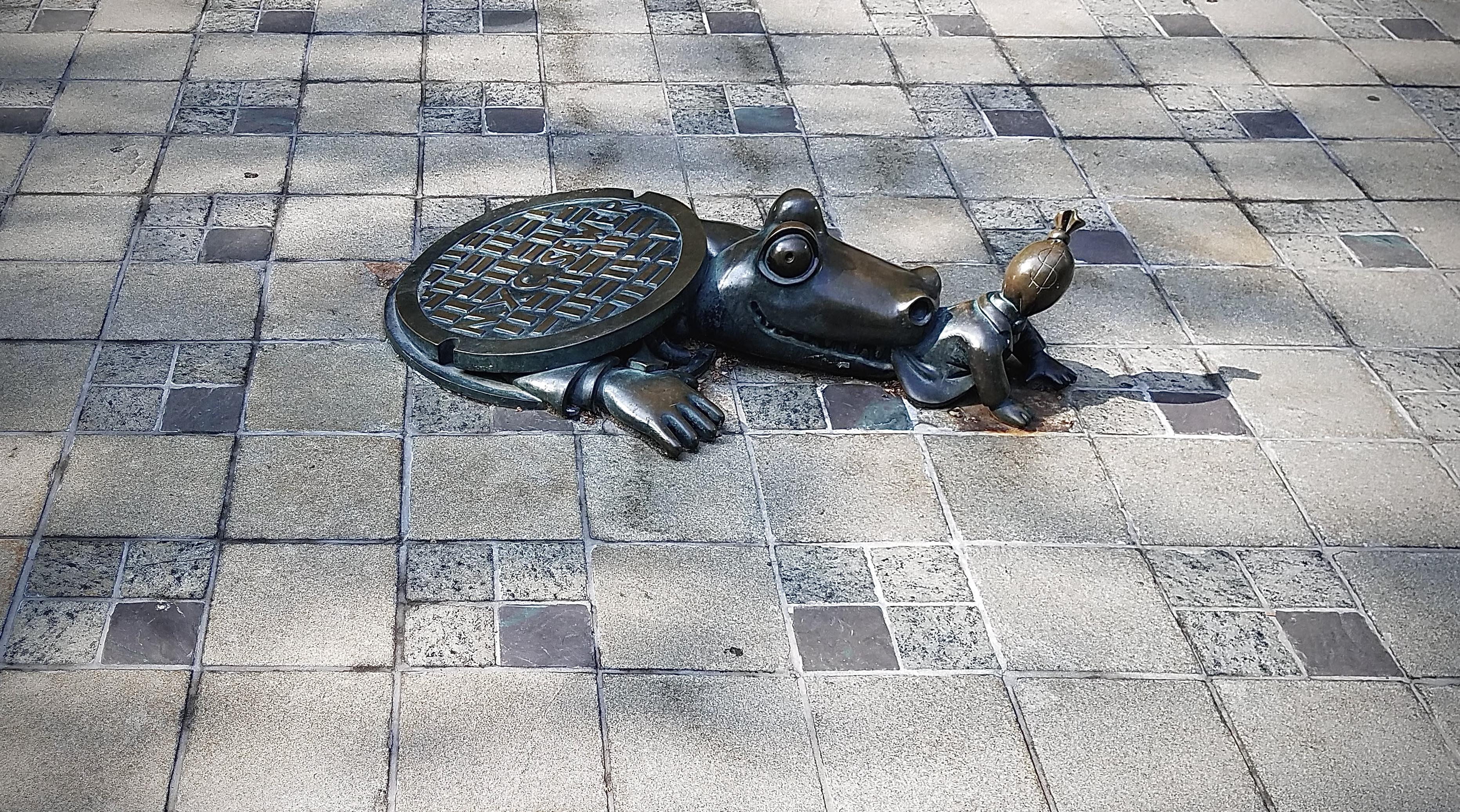A color, wide-angle photograph in landscape orientation that captures a bronze sculpture, part of Tom Otterness's "Life Underground" series, features an alligator emerging from a sewer. The creature’s head and one arm are visible as if it is crawling out from beneath a circular manhole cover, which sits directly on its back like a shell. The alligator has large, bulging eyes and a wide, mischievous expression. It has grabbed a small, humanoid figure by the waist with its jaws; the figure is depicted with a money bag for a head, complete with a dollar sign, emphasizing the artist's play on themes of capitalism and urban folklore. The installation is set against a ground of checkered stone pavers in alternating shades of light grey and tan. The bronze statue has a dark, weathered patina that contrasts with the neutral tones of the plaza floor. Shadows are cast by the sculpture from the afternoon sun, which highlights the smooth, rounded contours of the alligator's snout and the textured ridges of the manhole cover.
