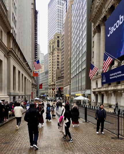 A color, wide-angle photograph in portrait orientation that captures a bustling pedestrian scene on Broad Street in the Financial District neighborhood of Manhattan, looking south from Wall Street. The ground is paved with glistening, wet cobblestones that reflect the overcast sky from recent light rain. On the right, the neoclassical architecture of the New York Stock Exchange dominates the foreground, draped with a massive blue corporate banner for "Jacobs" being listed, and two prominent American flags. To the left, the heavy stone facade of Federal Hall creates a narrow urban canyon effect. A black metal fence runs along the right side of the walkway, separating the main pedestrian path from the building's entrance. The street is filled with people dressed in layered winter clothing, including heavy coats, scarves, and beanies. In the immediate foreground, several pedestrians walk away from the camera, including a man in a black puffer jacket and a woman carrying a tan coat. On the right, a man in a dark jacket and grey vest stands near the fence, looking toward the camera. In the background, the dense architecture of the Financial District rises into a misty sky, with a mix of historic stone buildings and modern glass skyscrapers. Another American flag and orange Hermès flag are visible, and few white security tents are visible further down the street, amidst the crowd of tourists and commuters navigating the street.