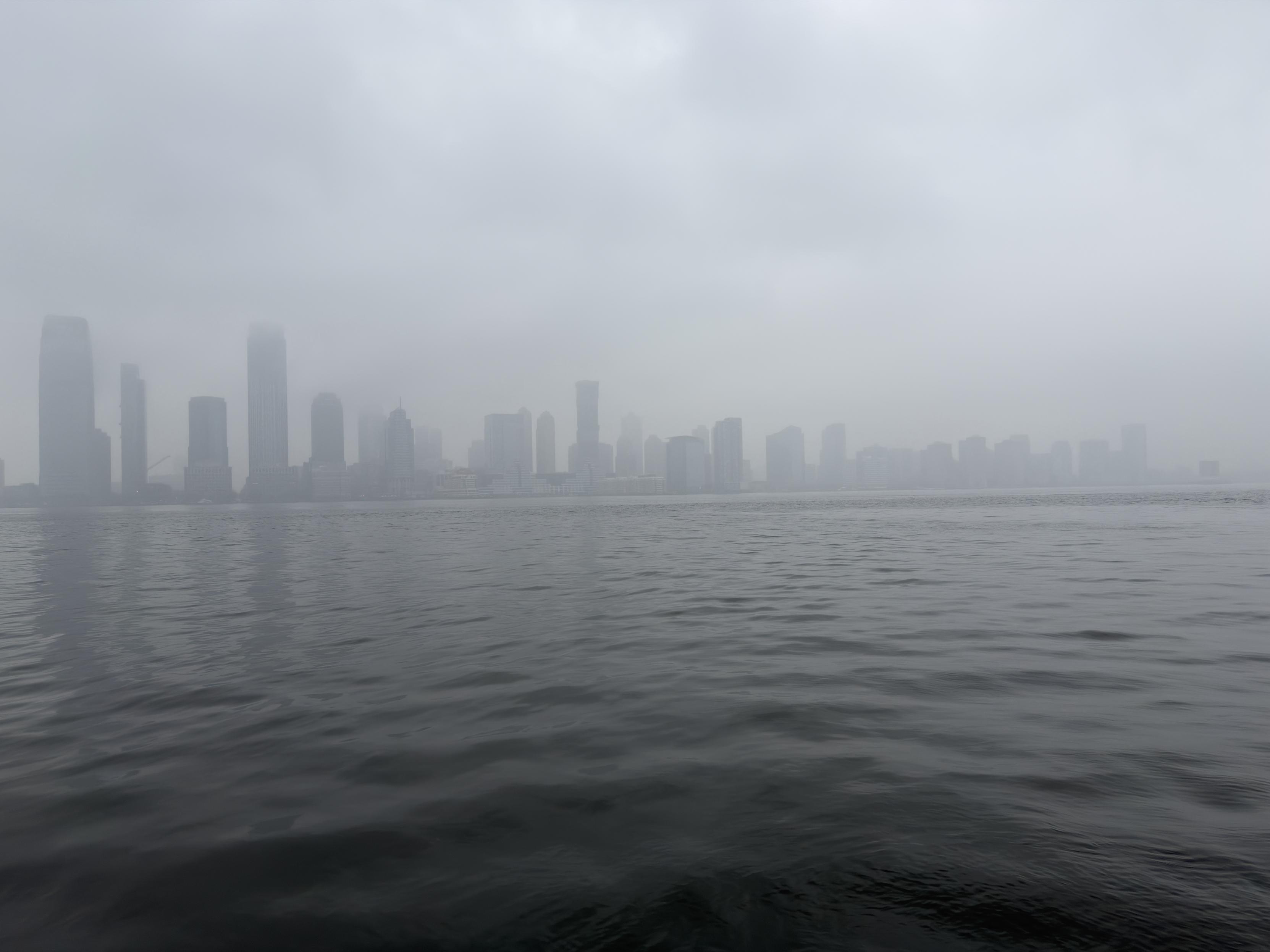 A color, wide-angle photograph in landscape orientation that captures a heavy, monochromatic view of the Jersey City skyline as seen from across the Hudson River in Manhattan. The composition is split nearly in half by a flat horizon line, with the dark, rippling water of the river in the foreground and a dense, overcast sky above. A thick blanket of grey fog hangs over the scene, partially obscuring the upper reaches of the skyscrapers and softening their architectural details into ghostly, desaturated, silhouetted shapes. The water is a deep, charcoal grey with small, rhythmic waves that create a textured surface reflecting the somber light of the sky. The skyline itself stretches across the middle ground, featuring a series of high-rise buildings in varying heights and widths. On the far left, the tallest structures stand as muted, dark pillars that disappear into the low-hanging clouds. As the eye moves to the right, the buildings become shorter, more distant and faint, eventually fading into a pale grey mist where the city meets the horizon. The overall atmosphere is quiet, cold, and ethereal, with the heavy atmospheric perspective stripping away most color and leaving only a range of tonal greys, faint greens, blues, and dark silhouettes.