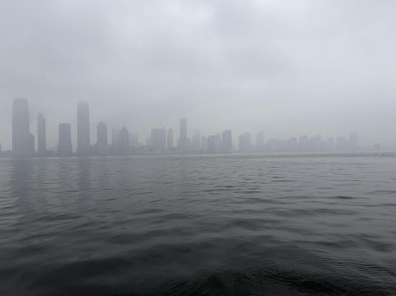 A color, wide-angle photograph in landscape orientation that captures a heavy, monochromatic view of the Jersey City skyline as seen from across the Hudson River in Manhattan. The composition is split nearly in half by a flat horizon line, with the dark, rippling water of the river in the foreground and a dense, overcast sky above. A thick blanket of grey fog hangs over the scene, partially obscuring the upper reaches of the skyscrapers and softening their architectural details into ghostly, desaturated, silhouetted shapes. The water is a deep, charcoal grey with small, rhythmic waves that create a textured surface reflecting the somber light of the sky. The skyline itself stretches across the middle ground, featuring a series of high-rise buildings in varying heights and widths. On the far left, the tallest structures stand as muted, dark pillars that disappear into the low-hanging clouds. As the eye moves to the right, the buildings become shorter, more distant and faint, eventually fading into a pale grey mist where the city meets the horizon. The overall atmosphere is quiet, cold, and ethereal, with the heavy atmospheric perspective stripping away most color and leaving only a range of tonal greys, faint greens, blues, and dark silhouettes.