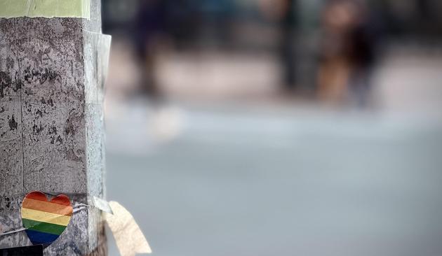 A color, medium-angle photograph in landscape orientation that captures a weathered, gray stone lamppost positioned on the left side of the frame. Attached to the lower portion of the pillar is a small, heart-shaped sticker featuring the classic six-color rainbow pride flag. The sticker is slightly creased and worn, secured to the rough surface with thin, clear adhesive that wraps partially around the post. Above the heart is a small, tattered piece of the bottom of a paper advertisement. Adjacent to the heart on the right edge of the lamppost is a small piece of curling paper. The background is heavily blurred, creating a soft bokeh effect that captures the urban atmosphere of the West Village in Manhattan. Muted tones of gray pavement and distant, indistinct shapes of pedestrians and city architecture provide a sense of place without distracting from the central symbol of the pride heart. The early evening lighting is soft and even, highlighting the gritty texture of the lamppost and dramatically emphasizing the pride heart.