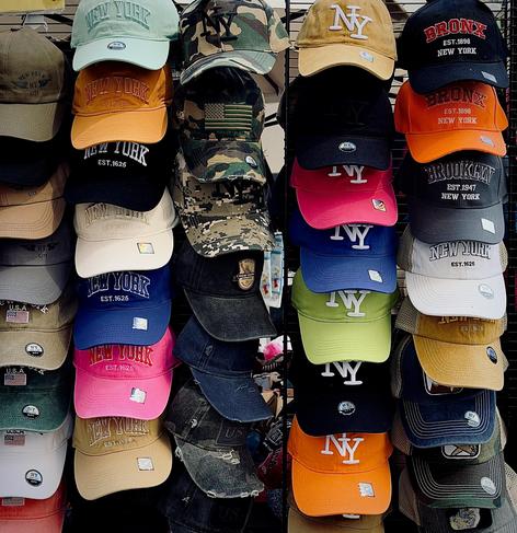A color, medium-angle photograph in square aspect ratio that captures the street scene along Eighth Avenue in the Garment District neighborhood of Manhattan. The focal point of the image is a dense vertical display which features dozens of baseball caps arranged in five distinct columns on a black wire rack next to a street vendor table. The collection showcases a variety of New York-themed designs, primarily focusing on "New York" arched text, "NY" interlocking logos, and specific borough names like "Bronx" and "Brooklyn." The color palette is expansive, ranging from neutral tones like olive green, khaki, and navy to vibrant neon greens, hot pinks, and bright oranges. Several hats feature camouflage patterns, and others incorporate small American flag patches or circular "NY City" authenticity stickers on the brims. The texture of the hats varies from smooth cotton twill to distressed, frayed brims and trucker-style mesh backing. The embroidery is prominent, with many hats using thick, raised white or contrast-colored stitching for the lettering. In the center-right column, a series of solid-colored caps features a large, white interlocking "NY" logo, stacked from top to bottom in tan, black, pink, blue, lime green, and orange. The overall arrangement is tightly packed, creating a repetitive but colorful grid.