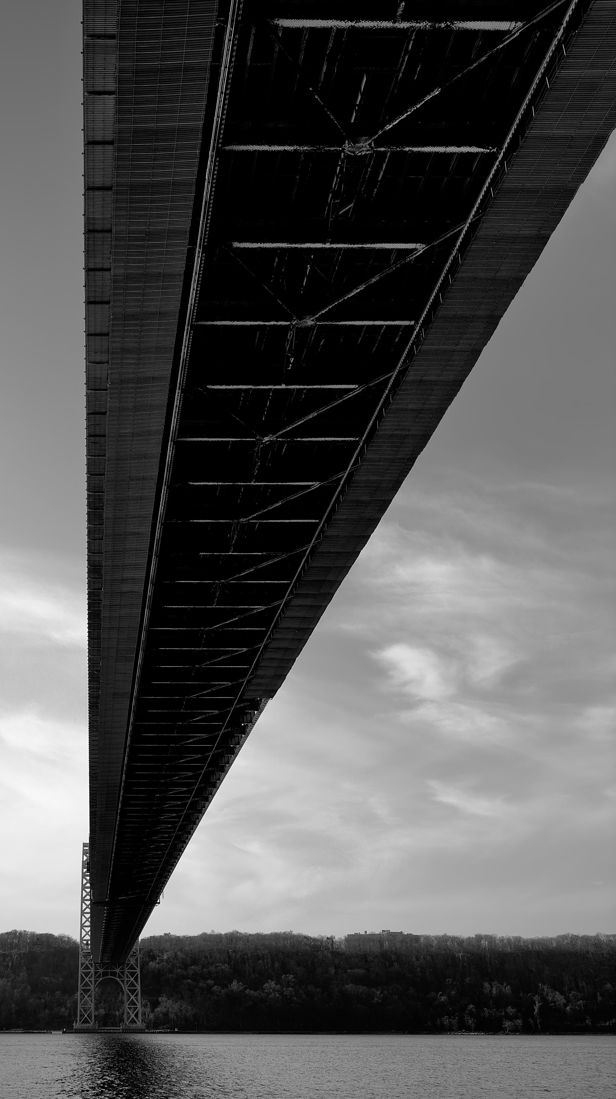A noir, wide-angle photograph in portrait orientation that captures a low-angle perspective from beneath the George Washington Bridge, looking across the Hudson River toward New Jersey. The composition is dominated by the massive, dark silhouette of the bridge deck, which cuts diagonally from the top right corner down toward the lower left. This angle emphasizes the geometry of the structure, revealing the intricate web of steel girders and diagonal bracing that support the roadway above. In the distance, the bridge’s steel lattice tower rises from the water, grounded on the far shore. Below the bridge, the dark, forested cliffs of New Jersey stretch across the horizon under a pale, overcast sky filled with wispy, elongated clouds. The water in the foreground is calm, textured with small ripples that catch the light, creating a shimmering contrast to the heavy, dark ironwork overhead. The overall mood is one of architectural scale and solitude, captured through a gritty, cinematic lens.
