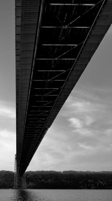 A noir, wide-angle photograph in portrait orientation that captures a low-angle perspective from beneath the George Washington Bridge, looking across the Hudson River toward New Jersey. The composition is dominated by the massive, dark silhouette of the bridge deck, which cuts diagonally from the top right corner down toward the lower left. This angle emphasizes the geometry of the structure, revealing the intricate web of steel girders and diagonal bracing that support the roadway above. In the distance, the bridge’s steel lattice tower rises from the water, grounded on the far shore. Below the bridge, the dark, forested cliffs of New Jersey stretch across the horizon under a pale, overcast sky filled with wispy, elongated clouds. The water in the foreground is calm, textured with small ripples that catch the light, creating a shimmering contrast to the heavy, dark ironwork overhead. The overall mood is one of architectural scale and solitude, captured through a gritty, cinematic lens.