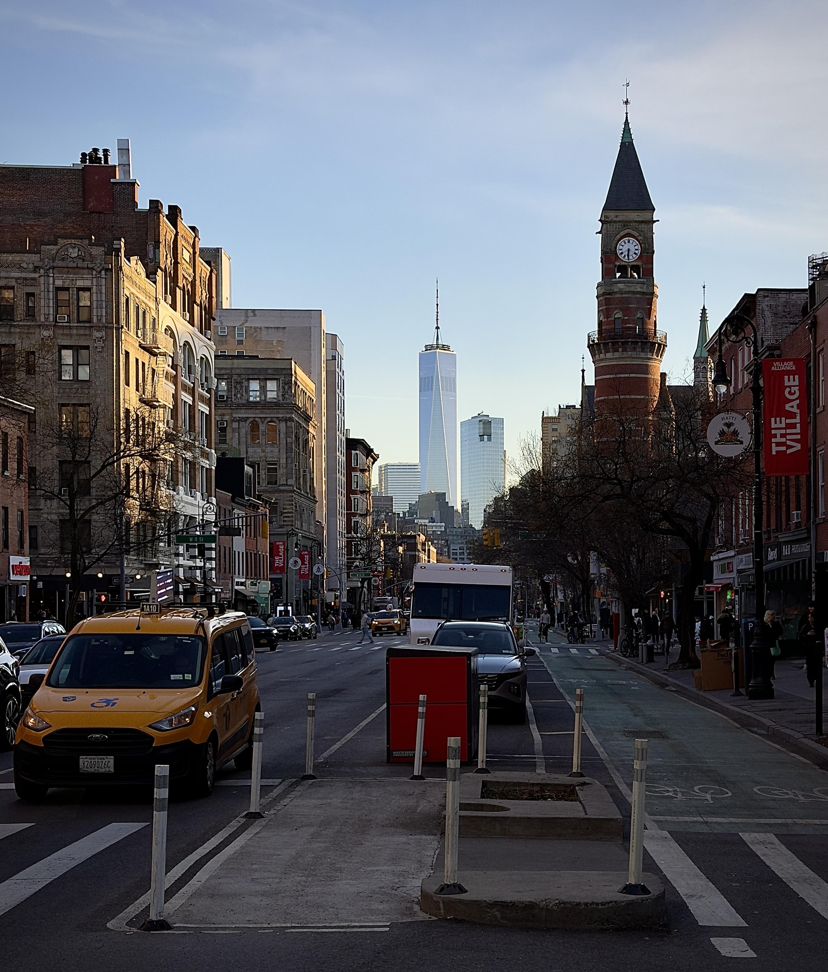 A color, wide-angle photograph in portrait orientation that captures the street scene along Sixth Avenue in the West Village neighborhood of Manhattan. In the foreground, a bright yellow Ford Transit taxi with accessibility decals is positioned on the left, while a dedicated green bike lane with white bicycle symbols runs along the right side of the road. On the right, the historic Jefferson Market Library stands out with its red brick facade and a prominent Victorian Gothic clock tower capped by a dark, pointed spire. A red "The Village" banner hangs from a nearby lamppost, and bare winter trees line the sidewalk where a few pedestrians are visible. The mid-ground is filled with city traffic, including several cars and a white truck, all moving toward the horizon. The street is flanked by traditional multi-story apartment buildings with fire escapes and storefronts on the left, and lower-profile commercial buildings on the right. In the far distance, the sleek, glass-clad One World Trade Center rises high above the surrounding skyline, its needle-like spire piercing a clear, pale blue sky. The lighting is typical of early evening, casting long, soft shadows and a warm glow over the architecture and the paved urban landscape.