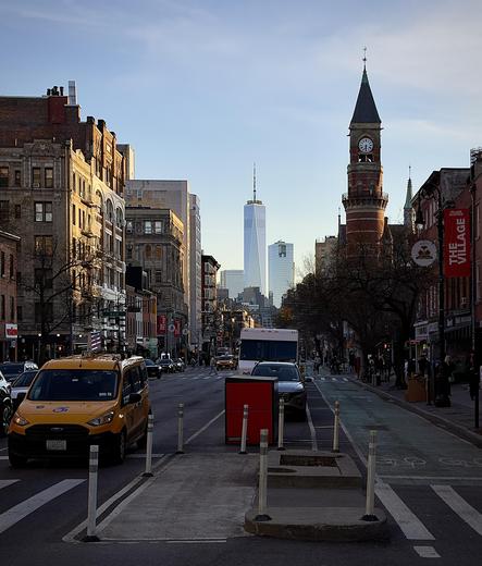 A color, wide-angle photograph in portrait orientation that captures the street scene along Sixth Avenue in the West Village neighborhood of Manhattan. In the foreground, a bright yellow Ford Transit taxi with accessibility decals is positioned on the left, while a dedicated green bike lane with white bicycle symbols runs along the right side of the road. On the right, the historic Jefferson Market Library stands out with its red brick facade and a prominent Victorian Gothic clock tower capped by a dark, pointed spire. A red "The Village" banner hangs from a nearby lamppost, and bare winter trees line the sidewalk where a few pedestrians are visible. The mid-ground is filled with city traffic, including several cars and a white truck, all moving toward the horizon. The street is flanked by traditional multi-story apartment buildings with fire escapes and storefronts on the left, and lower-profile commercial buildings on the right. In the far distance, the sleek, glass-clad One World Trade Center rises high above the surrounding skyline, its needle-like spire piercing a clear, pale blue sky. The lighting is typical of early evening, casting long, soft shadows and a warm glow over the architecture and the paved urban landscape.