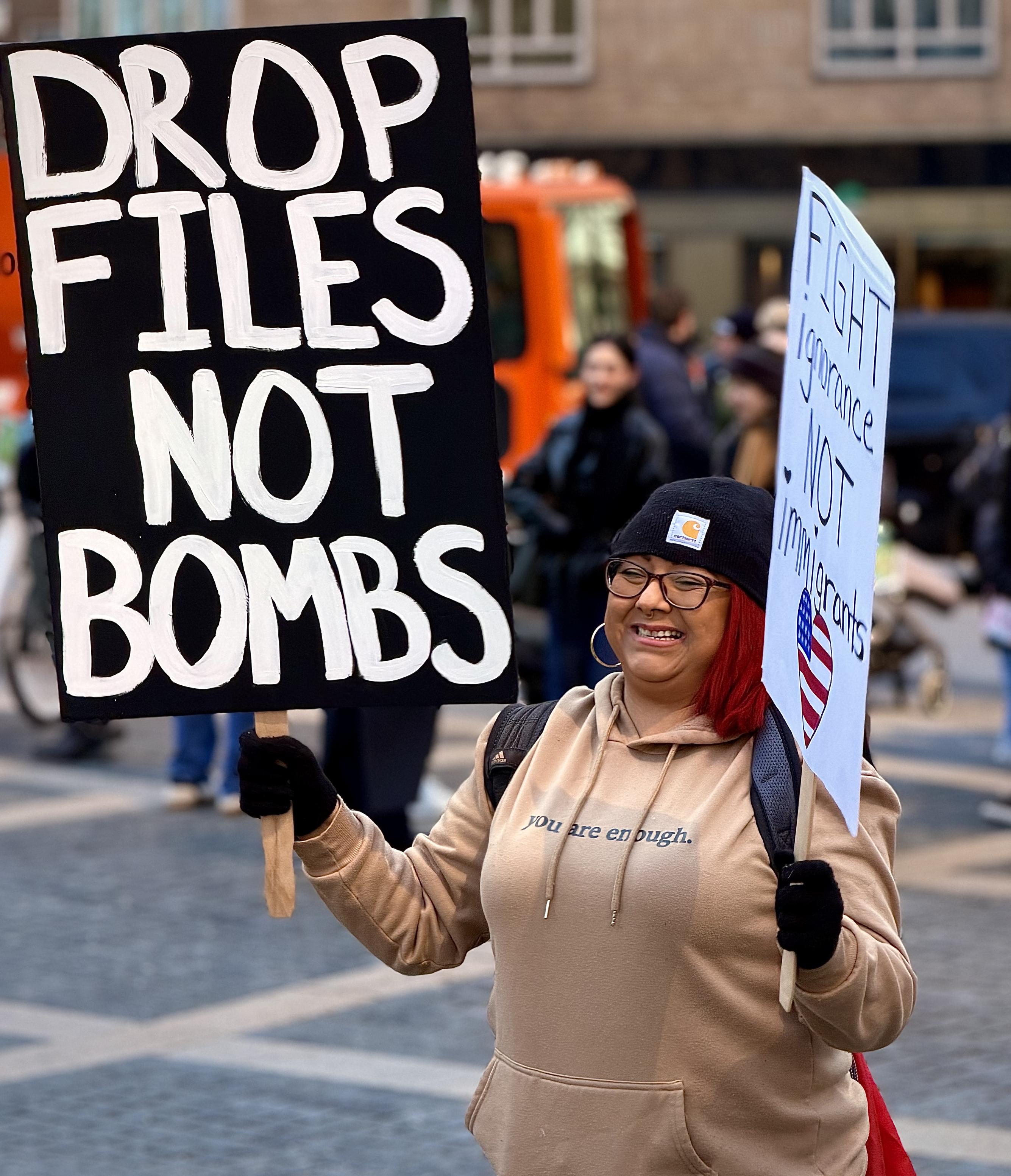 A color, medium-angle photograph in portrait orientation that captures a protestor standing in Columbus Circle. A woman with vibrant red hair, wearing a black beanie with a Carhartt logo, dark-rimmed glasses, and a tan hoodie that reads "you are enough." In her right hand, she holds a large black sign with bold white hand-painted letters that read "DROP FILES NOT BOMBS." In her left hand, she carries a smaller white sign that says "FIGHT ignorance NOT immigrants," featuring a heart-shaped American flag icon. The background shows a bustling outdoor city environment with cobblestone ground and other individuals blurred in the distance. To the left, a bright orange bus is partially visible, and several people in winter clothing walk through the plaza. the lighting is bright and natural, capturing the protestor in a sharp focus that highlights the texture of the signs and the cheerful expression on her face amidst the active demonstration. Portrait shared with permission, the subject is Sandra from Mott Haven, the Bronx.
