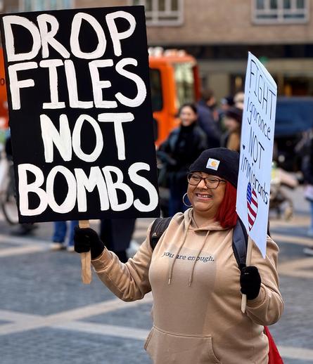 A color, medium-angle photograph in portrait orientation that captures a protestor standing in Columbus Circle. A woman with vibrant red hair, wearing a black beanie with a Carhartt logo, dark-rimmed glasses, and a tan hoodie that reads "you are enough." In her right hand, she holds a large black sign with bold white hand-painted letters that read "DROP FILES NOT BOMBS." In her left hand, she carries a smaller white sign that says "FIGHT ignorance NOT immigrants," featuring a heart-shaped American flag icon. The background shows a bustling outdoor city environment with cobblestone ground and other individuals blurred in the distance. To the left, a bright orange bus is partially visible, and several people in winter clothing walk through the plaza. the lighting is bright and natural, capturing the protestor in a sharp focus that highlights the texture of the signs and the cheerful expression on her face amidst the active demonstration. Portrait shared with permission, the subject is Sandra from Mott Haven, the Bronx.