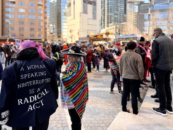 A color, wide-angle photograph in landscape orientation that captures a No Kings outdoor protest in Columbus Circle, Manhattan. The scene is surrounded by tall modern buildings under a bright, clear sky. In the foreground, the primary focal point is a woman seen from behind with vibrant pink hair tied in a ponytail. She is wearing a dark navy blazer featuring a bold, white-lettered political statement that reads: “A WOMAN SPEAKING UNINTERRUPTED IS A RADICAL ACT.” On the sleeve is reads “Fuck Trump.” Just beside her, another individual stands in profile wearing a wide-brimmed black sombrero with silver trim and a multi-colored striped serape, adding to the eclectic and expressive nature of the crowd. The midground and background are filled with a diverse group of demonstrators and onlookers, many in winter coats and hats. To the right, a man in a tan jacket and another in a black puffy vest stand near stone steps, while further back, yellow umbrellas from street vendor carts and various colorful signs are visible among the throng of people. The atmosphere appears active and densely populated, framed by the towering glass and brick architecture of the midtown skyline.
