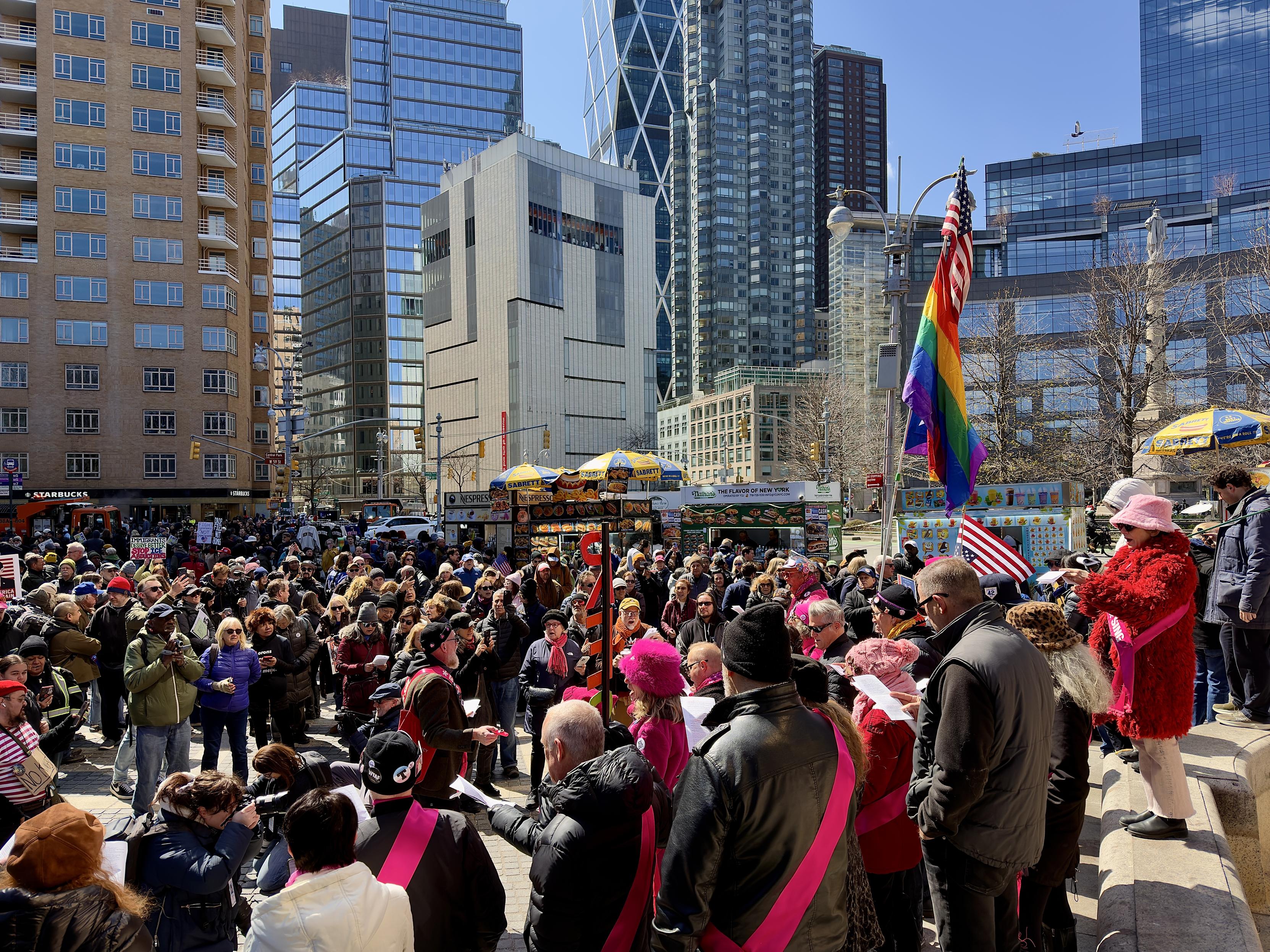 A color, wide-angle photograph in landscape orientation that captures a vibrant and crowded protest scene at Columbus Circle in Manhattan, set against a backdrop of towering modern glass skyscrapers and historic brick buildings. A dense, diverse crowd of people occupies the plaza. In the foreground, several individuals are holding sheets of paper while singing together. A tall flagpole stands prominently on the right, flying a large rainbow Pride flag topped with a smaller American flag, while a few smaller American flags and signs are scattered throughout the crowd. The atmosphere is energetic, illuminated by bright, direct early afternoon sunlight that creates sharp shadows across the pavement. The middle ground is filled with typical New York City street elements, including several Sabrett hot dog carts with their iconic blue and yellow umbrellas and a green food truck labeled "The Flavor of New York." Demonstrators hold various signs, with one visible in the distance referencing immigrant rights. To the far left, a Starbucks storefront is partially visible at the base of a brown residential building. On the right, a person in a striking fuzzy red coat and pink hat stands on a stone ledge, overlooking the gathering. The architecture in the background features the distinct geometric, crisscrossed bracing of the Hearst Tower, grounding the scene in the heart of Midtown Manhattan.
