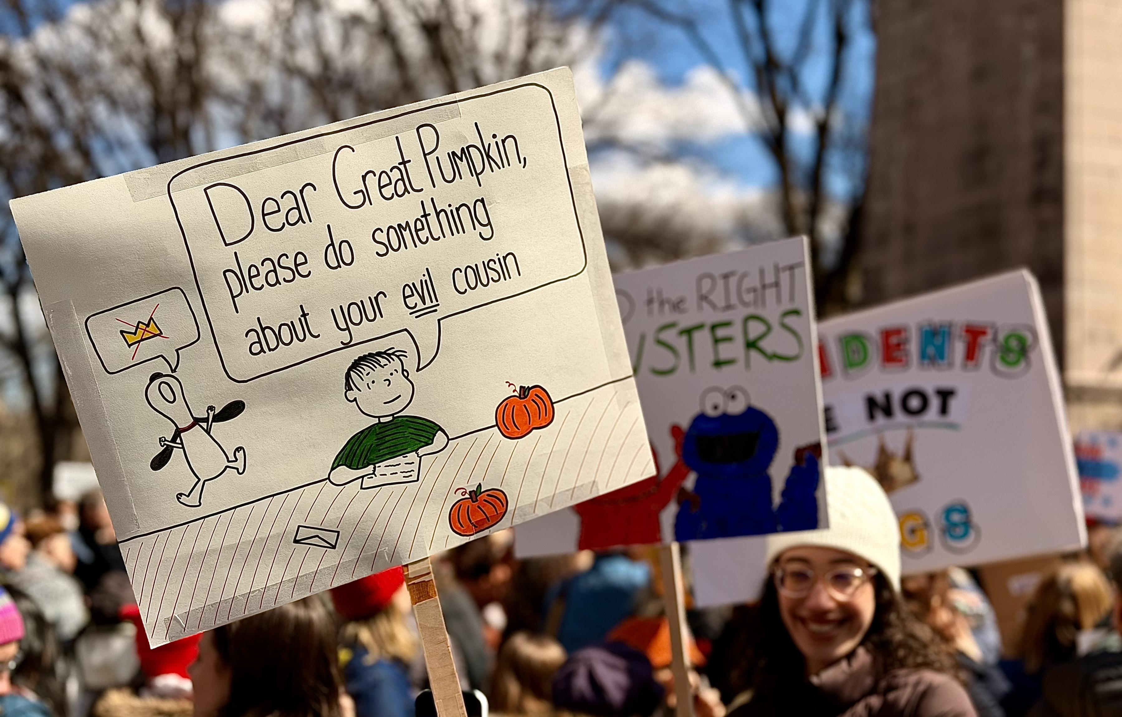 A color, medium-angle photograph that captures the No Kings protest in Columbus Circle, Manhattan. There is a protestor holding a large, hand-drawn sign featuring characters from the Peanuts comic strip. The sign depicts Linus sitting at a desk with two pumpkins, writing a letter that appears in a large speech bubble above him. The text reads, "Dear Great Pumpkin, please do something about your evil cousin." Beside him, a line drawing of Snoopy is shown with a smaller thought bubble containing a golden crown with a red "X" struck through it, signaling an anti-monarchy sentiment. The sign is held aloft by a wooden stake, positioned prominently in the foreground against a blurred background of other demonstrators and bare winter trees. To the right, a smiling woman in a white beanie and glasses holds another sign featuring the Sesame Street characters Elmo and Cookie Monster. In the far background, a third sign is partially visible with colorful block lettering that reads "Presidents are not kings" above a crown icon. The lighting is bright and natural, suggesting a clear day, and the crowd is dressed in heavy winter coats and hats.