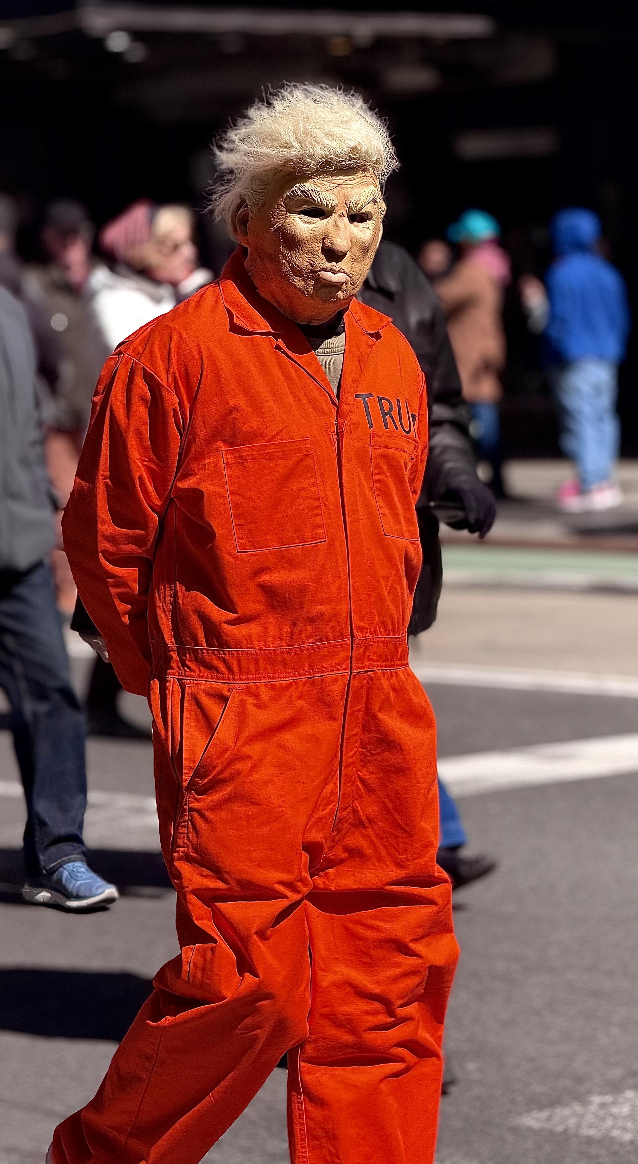 A color, medium-angle photograph in portrait orientation that captures a person standing at the front of the No Kings protest, wearing a full-body orange prison-style jumpsuit and a rubber mask portraying Donald Trump. The mask has a caricature-like quality with deep wrinkles, pursed lips, and a prominent tuft of messy, bright blonde hair. On the left chest area of the jumpsuit, the letters "TRU" are visible, suggesting the name "TRUMP" continues behind the fold of the fabric. The person is positioned with their body angled slightly toward the left while their head is turned forward, standing with their hands held behind their back in a pose resembling a prisoner. The background is a blurred Times Square with several pedestrians walking in various directions. The sunlight is bright, creating sharp shadows on the person’s orange jumpsuit and highlighting the textured surface of the mask. A green bike lane and a white crosswalk stripe are visible on the asphalt ground, while people in the distance wear winter clothing like hoodies and jackets. The focus remains sharply on the individual in the costume, emphasizing the political satire of the attire within the context of a public demonstration.