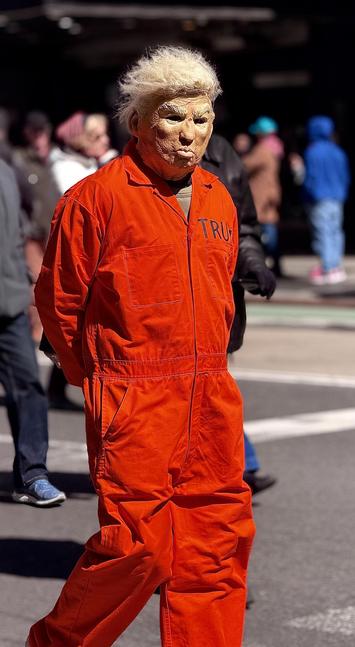 A color, medium-angle photograph in portrait orientation that captures a person standing at the front of the No Kings protest, wearing a full-body orange prison-style jumpsuit and a rubber mask portraying Donald Trump. The mask has a caricature-like quality with deep wrinkles, pursed lips, and a prominent tuft of messy, bright blonde hair. On the left chest area of the jumpsuit, the letters "TRU" are visible, suggesting the name "TRUMP" continues behind the fold of the fabric. The person is positioned with their body angled slightly toward the left while their head is turned forward, standing with their hands held behind their back in a pose resembling a prisoner. The background is a blurred Times Square with several pedestrians walking in various directions. The sunlight is bright, creating sharp shadows on the person’s orange jumpsuit and highlighting the textured surface of the mask. A green bike lane and a white crosswalk stripe are visible on the asphalt ground, while people in the distance wear winter clothing like hoodies and jackets. The focus remains sharply on the individual in the costume, emphasizing the political satire of the attire within the context of a public demonstration.