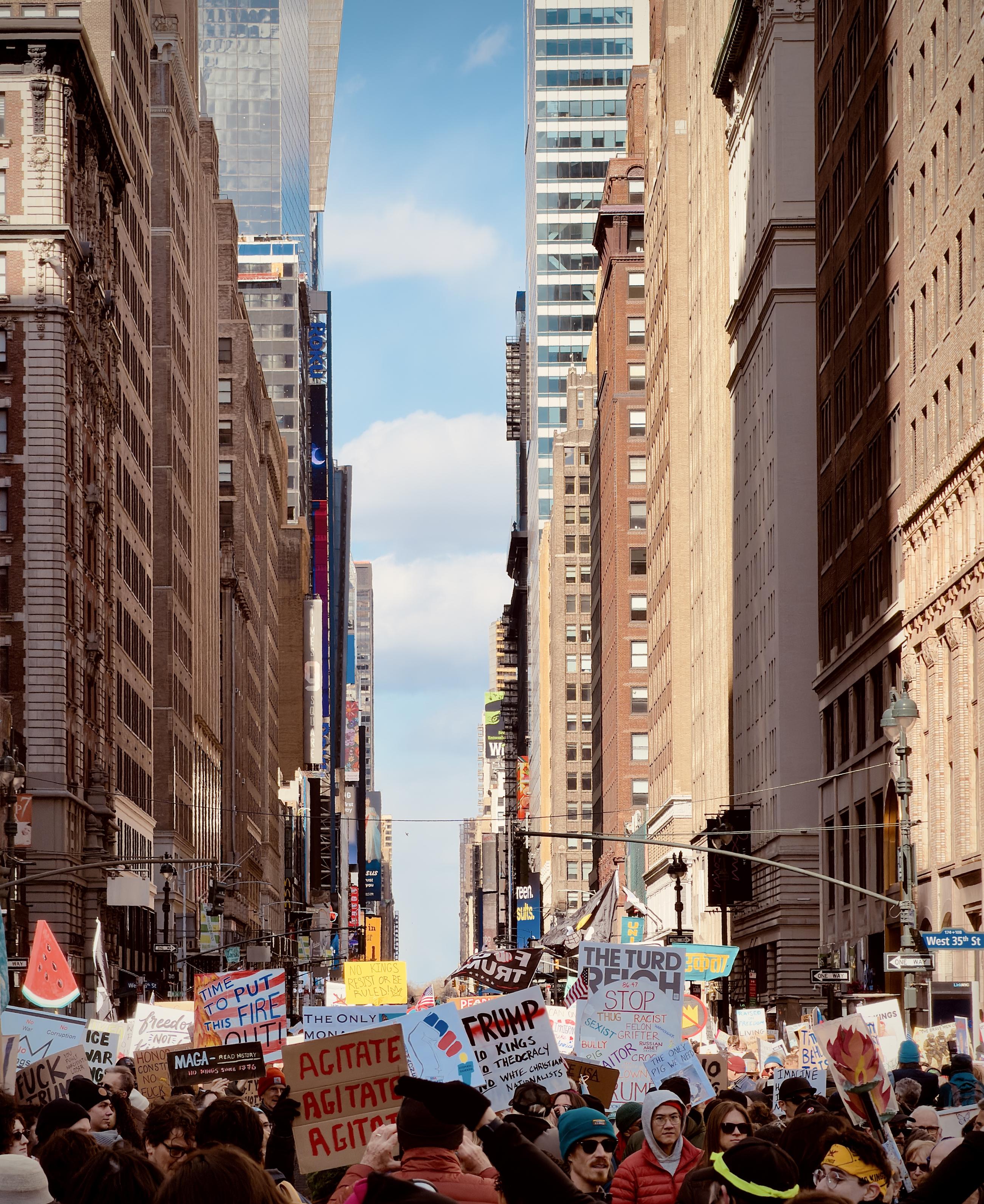 A color, wide-angle photograph in portrait orientation that captures a dense crowd of No Kings protesters marching down a narrow canyon of skyscrapers on 7th Avenue in the Chelsea neighborhood of Manhattan. The perspective is from within the crowd, looking north toward a sliver of blue sky framed by the towering architecture of the city. A street sign in the lower right corner identifies the intersection as West 35th Street. Protesters are dressed in winter clothing, including beanies and heavy jackets, and are carrying a wide variety of handmade signs. The signs feature bold text and political slogans such as "AGITATE AGITATE AGITATE," "TIME TO PUT THIS FIRE OUT!," and "NO KINGS," alongside various anti-Trump imagery and symbols like a large watermelon slice cutout. The composition emphasizes the massive scale of the march, with the sea of people stretching far into the distance between the sunlit and shadowed facades of the buildings. The lighting creates a high-contrast environment, with bright sunlight hitting the upper stories of the skyscrapers on the right while the street level remains in partial shadow. In the foreground, the backs of protesters' heads and the tops of their colorful signs dominate the frame, creating a sense of being physically present within the demonstration. The overall atmosphere is one of active civic engagement and urban movement.