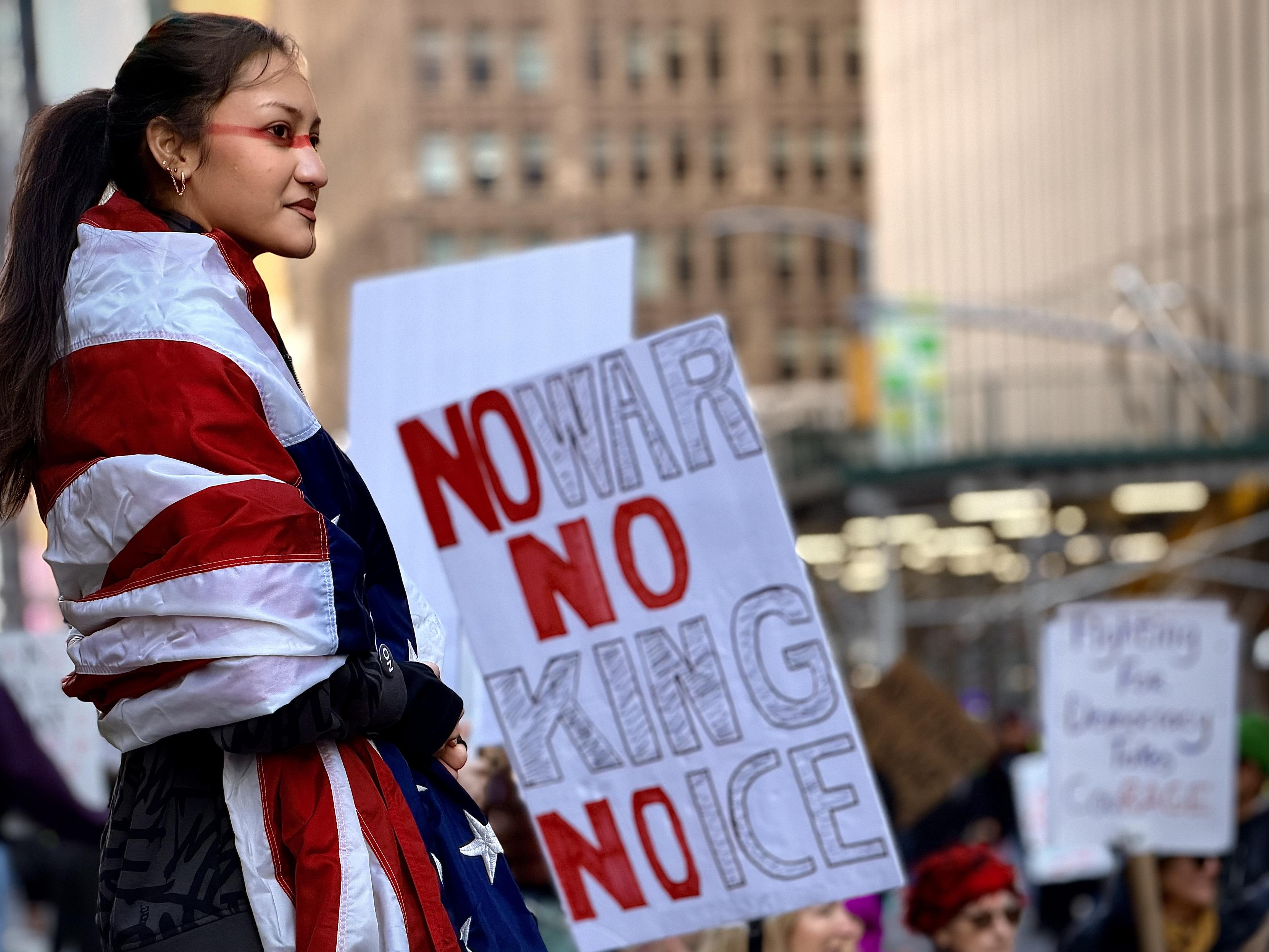 A color, wide-angle photograph in landscape orientation that captures a powerful moment during the No Kings protest along 7th Avenue in Manhattan. A young woman with dark hair pulled back into a ponytail stands in profile, looking off to the side with a determined and calm expression. She has a striking red horizontal line painted across her face, spanning from one cheekbone across the bridge of her nose to the other. She is wrapped in a large American flag, which is draped over her shoulders and torso like a shawl, the red and white stripes and blue field with white stars prominently visible. Positioned next to her is a large, hand-drawn protest sign someone else is holding that reads "NO WAR NO KING NO ICE" in bold, vertical lettering. The words "NO" are filled in with solid red, while "WAR," "KING," and "ICE" are rendered in large, hollow, outlined letters. In the blurred background, other protesters fill the street and city buildings create a sense of a large-scale public demonstration. The afternoon lighting is natural and soft, highlighting the woman as the central figure of the composition against the bustling urban environment.