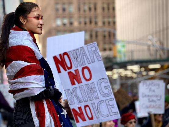 A color, wide-angle photograph in landscape orientation that captures a powerful moment during the No Kings protest along 7th Avenue in Manhattan. A young woman with dark hair pulled back into a ponytail stands in profile, looking off to the side with a determined and calm expression. She has a striking red horizontal line painted across her face, spanning from one cheekbone across the bridge of her nose to the other. She is wrapped in a large American flag, which is draped over her shoulders and torso like a shawl, the red and white stripes and blue field with white stars prominently visible. Positioned next to her is a large, hand-drawn protest sign someone else is holding that reads "NO WAR NO KING NO ICE" in bold, vertical lettering. The words "NO" are filled in with solid red, while "WAR," "KING," and "ICE" are rendered in large, hollow, outlined letters. In the blurred background, other protesters fill the street and city buildings create a sense of a large-scale public demonstration. The afternoon lighting is natural and soft, highlighting the woman as the central figure of the composition against the bustling urban environment.