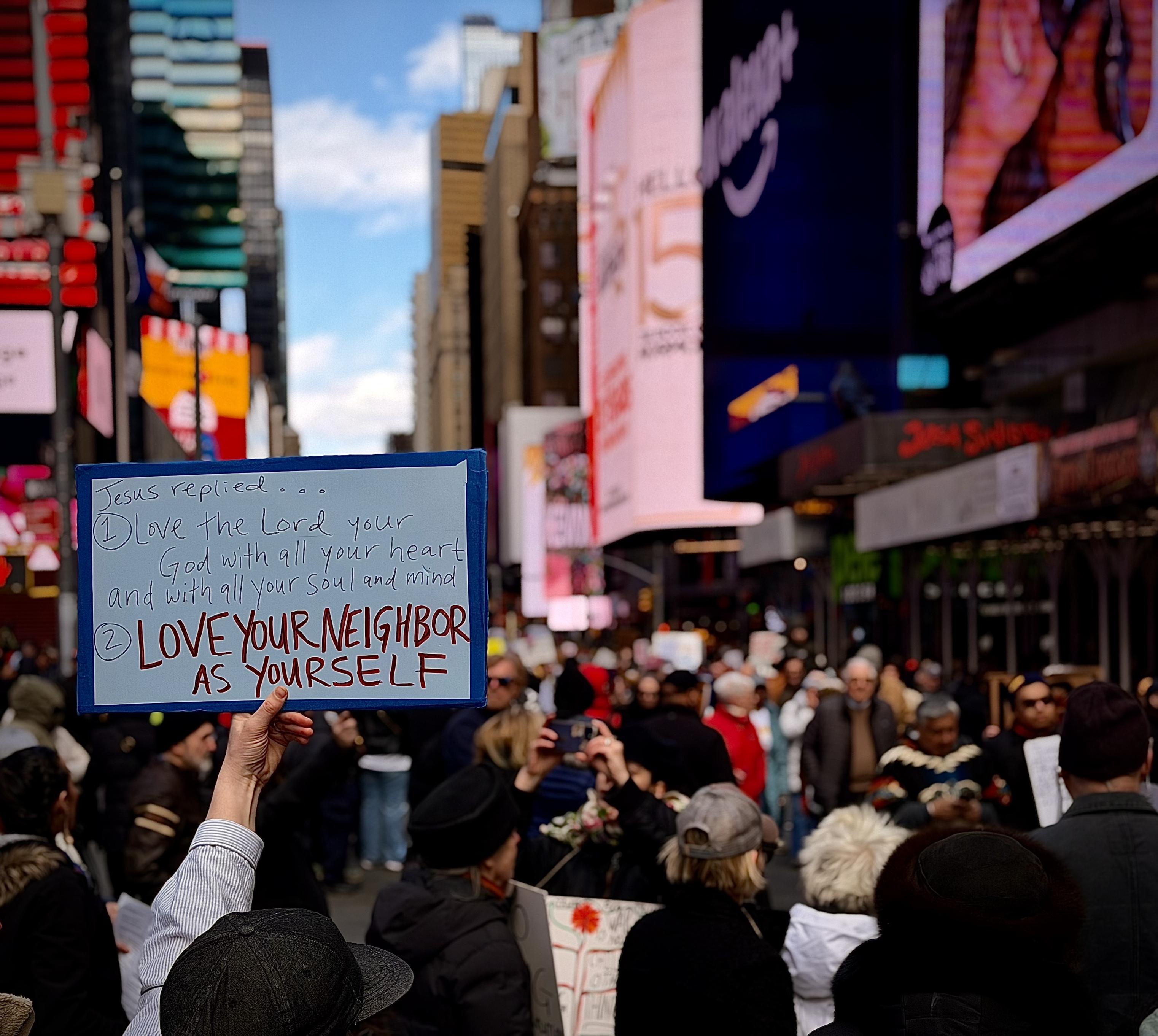 A color, wide-angle photograph in square aspect ratio that captures a dense crowd of demonstrators during the No Kings protest as it passed through Times Square, Manhattan. The perspective is from the plaza between Broadway and Seventh Avenue, looking northeast toward the iconic glowing billboards and architectural canyons of Midtown. In the foreground, a protestor’s arm reaches upward, holding a hand-painted blue and white sign that quotes religious scripture: "Jesus replied... 1) Love the Lord your God with all your heart and with all your soul and mind. 2) LOVE YOUR NEIGHBOR AS YOURSELF," with the final line emphasized in bold red lettering. The surrounding crowd is a diverse mix of individuals in winter attire, including beanies and heavy jackets, reflecting the brisk spring day. The background is a vibrant, shallow-focus blur of Times Square’s signature commercial displays, featuring large digital screens with pink and blue hues. The street is packed with people extending deep into the distance, framed by the scaffolding of New York City construction and the glass facades of skyscrapers under a bright, partly cloudy sky.