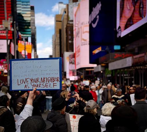 A color, wide-angle photograph in square aspect ratio that captures a dense crowd of demonstrators during the No Kings protest as it passed through Times Square, Manhattan. The perspective is from the plaza between Broadway and Seventh Avenue, looking northeast toward the iconic glowing billboards and architectural canyons of Midtown. In the foreground, a protestor’s arm reaches upward, holding a hand-painted blue and white sign that quotes religious scripture: "Jesus replied... 1) Love the Lord your God with all your heart and with all your soul and mind. 2) LOVE YOUR NEIGHBOR AS YOURSELF," with the final line emphasized in bold red lettering. The surrounding crowd is a diverse mix of individuals in winter attire, including beanies and heavy jackets, reflecting the brisk spring day. The background is a vibrant, shallow-focus blur of Times Square’s signature commercial displays, featuring large digital screens with pink and blue hues. The street is packed with people extending deep into the distance, framed by the scaffolding of New York City construction and the glass facades of skyscrapers under a bright, partly cloudy sky.