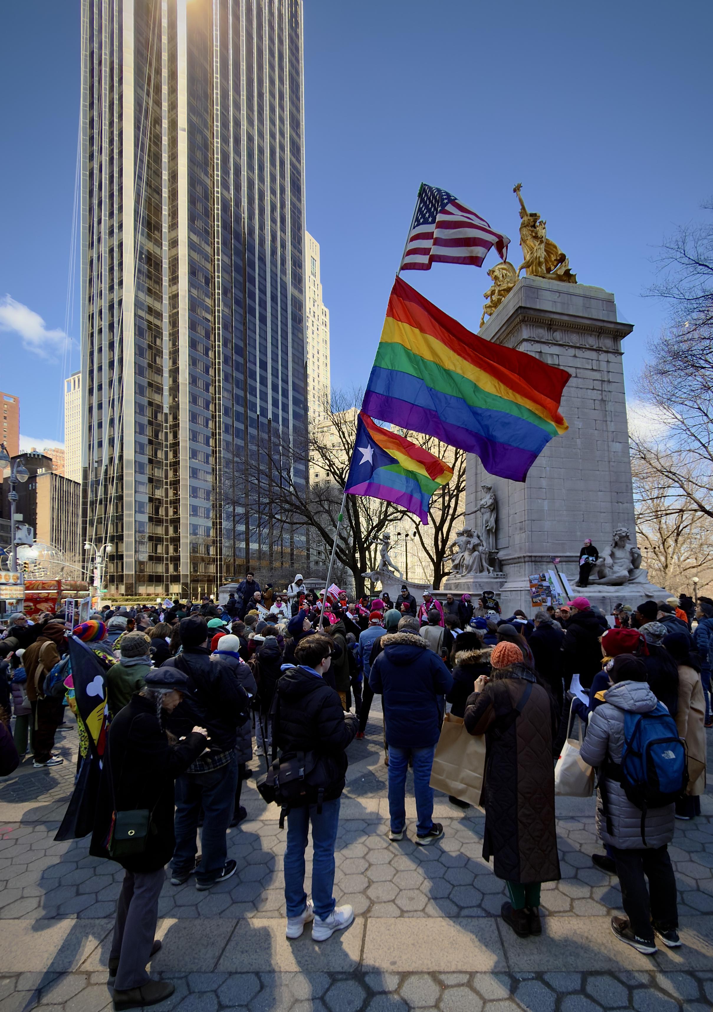 A color, wide-angle photograph in portrait orientation that captures Columbus Circle at Central Park West in Manhattan during the No Kings protest on March 28, 2026. In the foreground, a dense crowd of demonstrators has gathered around the base of the USS Maine National Monument at the Merchants' Gate entrance to Central Park. Protesters are seen holding a variety of handmade signs and banners with slogans such as "No Kings," "Defend Democracy," and "End the War," creating a vibrant tapestry of color against the grey stone of the monument’s pylon and its allegorical sculptures. A person is holding a flag pole with American and Pride flags. Dominating the background and providing a sharp ironic contrast to the human activity below is the Trump International Hotel & Tower at 1 Central Park West. The 52-story skyscraper's bronze-tinted glass facade is brilliantly illuminated by the morning sun, reflecting a shimmering golden glow that washes over the circle making the building appear as a massive, glowing monolith overlooking the protest.
