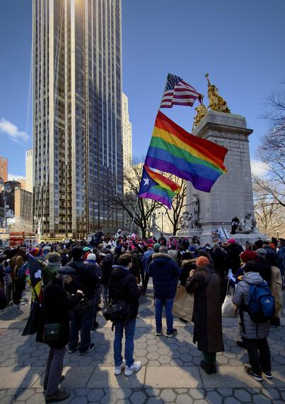 A color, wide-angle photograph in portrait orientation that captures Columbus Circle at Central Park West in Manhattan during the No Kings protest on March 28, 2026. In the foreground, a dense crowd of demonstrators has gathered around the base of the USS Maine National Monument at the Merchants' Gate entrance to Central Park. Protesters are seen holding a variety of handmade signs and banners with slogans such as "No Kings," "Defend Democracy," and "End the War," creating a vibrant tapestry of color against the grey stone of the monument’s pylon and its allegorical sculptures. A person is holding a flag pole with American and Pride flags. Dominating the background and providing a sharp ironic contrast to the human activity below is the Trump International Hotel & Tower at 1 Central Park West. The 52-story skyscraper's bronze-tinted glass facade is brilliantly illuminated by the morning sun, reflecting a shimmering golden glow that washes over the circle making the building appear as a massive, glowing monolith overlooking the protest.