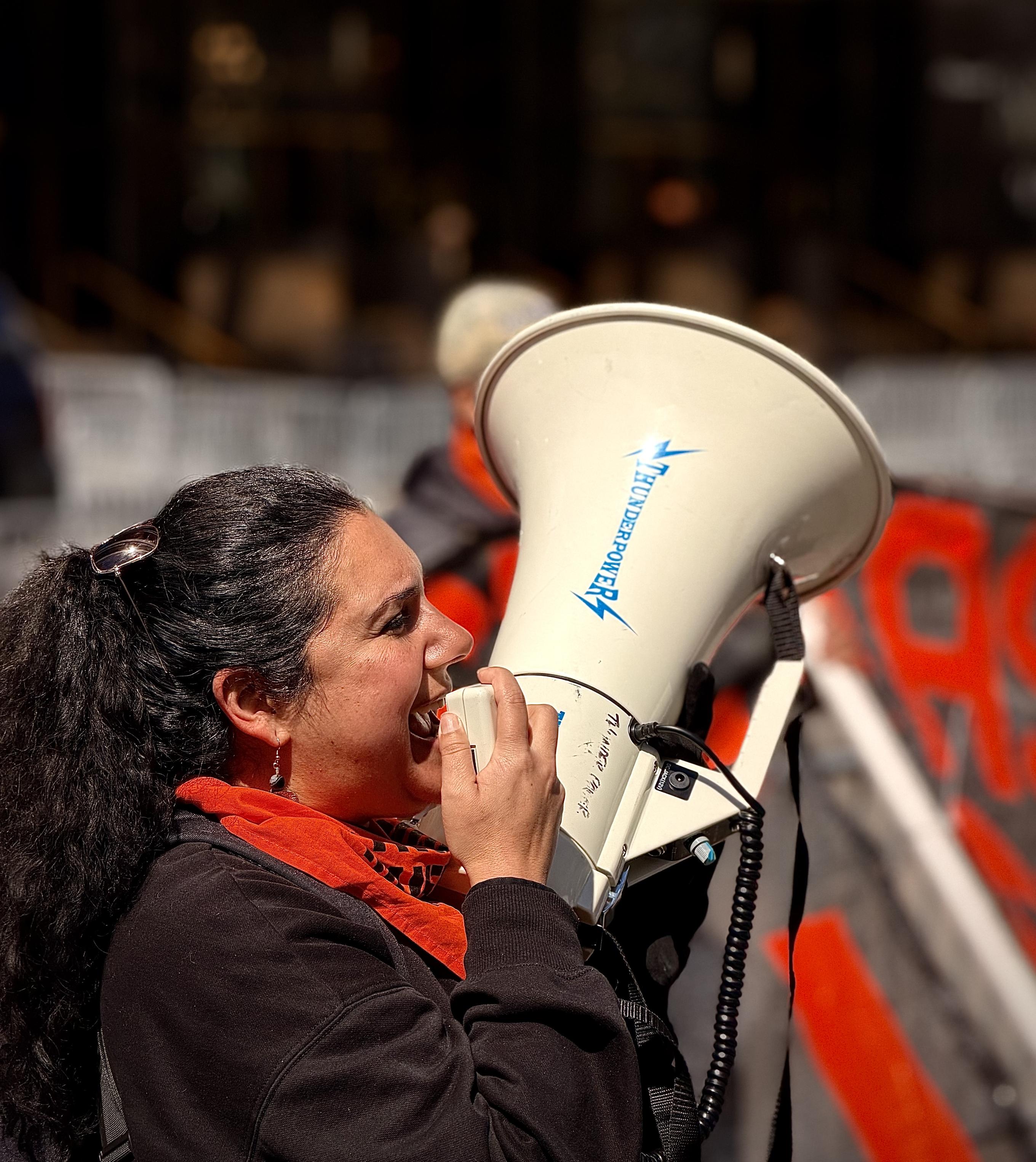 A color, wide-angle photograph in square aspect ratio that captures captures an activist in the midst of a "Resist Fascism" protest, positioned directly in front of the Trump International Hotel & Tower at 1 Central Park West in Manhattan. The woman, with her dark hair pulled back into a high ponytail, is shown in profile, her face expressive with passion as she leads a chant through a large, white ThunderPower bullhorn. She is dressed in a black hoodie and a vibrant red-and-black patterned scarf, a color scheme often associated with anti-fascist movements. The background of the image provides a sense of the urban protest environment, with the distinctive bronze-tinted glass of the Trump hotel looming behind her. Metal police barricades and a flash of a red protest banner are visible in the shallow depth of field, centering the focus entirely on her leadership. The lighting is bright and direct, highlighting the intensity of the moment and the grit of the demonstration as she rallies the crowd against the backdrop of one of the city's most controversial landmarks.