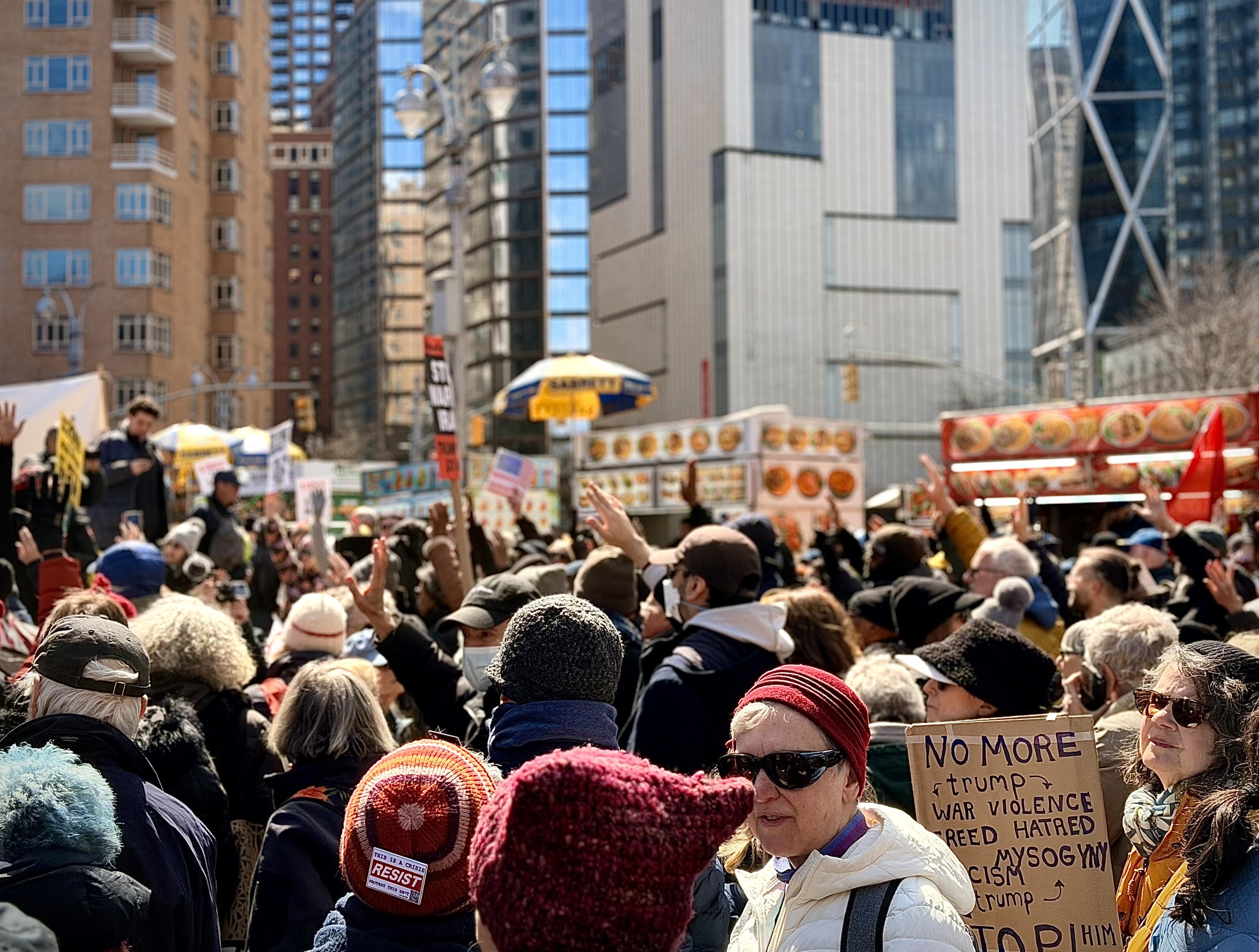 A color, wide-angle photograph in landscape orientation that captures an eye-level view of the dense crowd of protesters gathered at Columbus Circle in Manhattan for a No Kings event. The scene is filled with people of diverse ages and backgrounds, many wearing winter hats, coats, and sunglasses. Several individuals have their hands raised in the air, while others hold signs and American flags. In the lower right foreground, a woman in a white jacket and red beanie stands near a cardboard sign that reads "NO MORE trump, WAR VIOLENCE, GREED HATRED, RACISM MISOGYNY, trump, STOP HIM." Another protester in the lower left wears a knit hat with a "RESIST" pin attached, it reads “This is a crisis. Protest this shit.” The background features the towering urban architecture of midtown Manhattan, including a modern glass building with a distinct silver X-brace structural design and a traditional brick apartment complex. A row of yellow and red food carts with vibrant food imagery lines the edge of the plaza, partially obscured by the crowd. A speaker is visible on a raised platform in the mid-ground to the left, addressing a group. The lighting is bright and clear, suggesting a sunny day, with a shallow depth of field that keeps the central protesters in focus while the city buildings and street lamps softly blur into the distance.