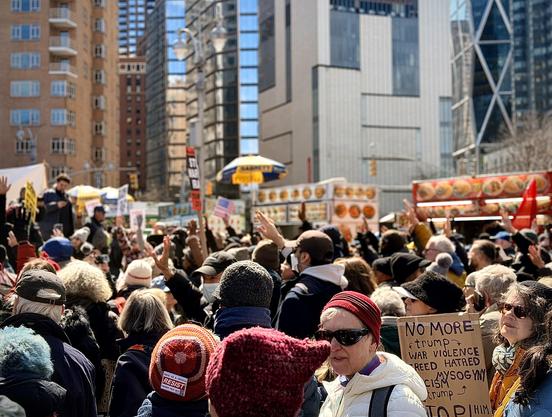 A color, wide-angle photograph in landscape orientation that captures an eye-level view of the dense crowd of protesters gathered at Columbus Circle in Manhattan for a No Kings event. The scene is filled with people of diverse ages and backgrounds, many wearing winter hats, coats, and sunglasses. Several individuals have their hands raised in the air, while others hold signs and American flags. In the lower right foreground, a woman in a white jacket and red beanie stands near a cardboard sign that reads "NO MORE trump, WAR VIOLENCE, GREED HATRED, RACISM MISOGYNY, trump, STOP HIM." Another protester in the lower left wears a knit hat with a "RESIST" pin attached, it reads “This is a crisis. Protest this shit.” The background features the towering urban architecture of midtown Manhattan, including a modern glass building with a distinct silver X-brace structural design and a traditional brick apartment complex. A row of yellow and red food carts with vibrant food imagery lines the edge of the plaza, partially obscured by the crowd. A speaker is visible on a raised platform in the mid-ground to the left, addressing a group. The lighting is bright and clear, suggesting a sunny day, with a shallow depth of field that keeps the central protesters in focus while the city buildings and street lamps softly blur into the distance.