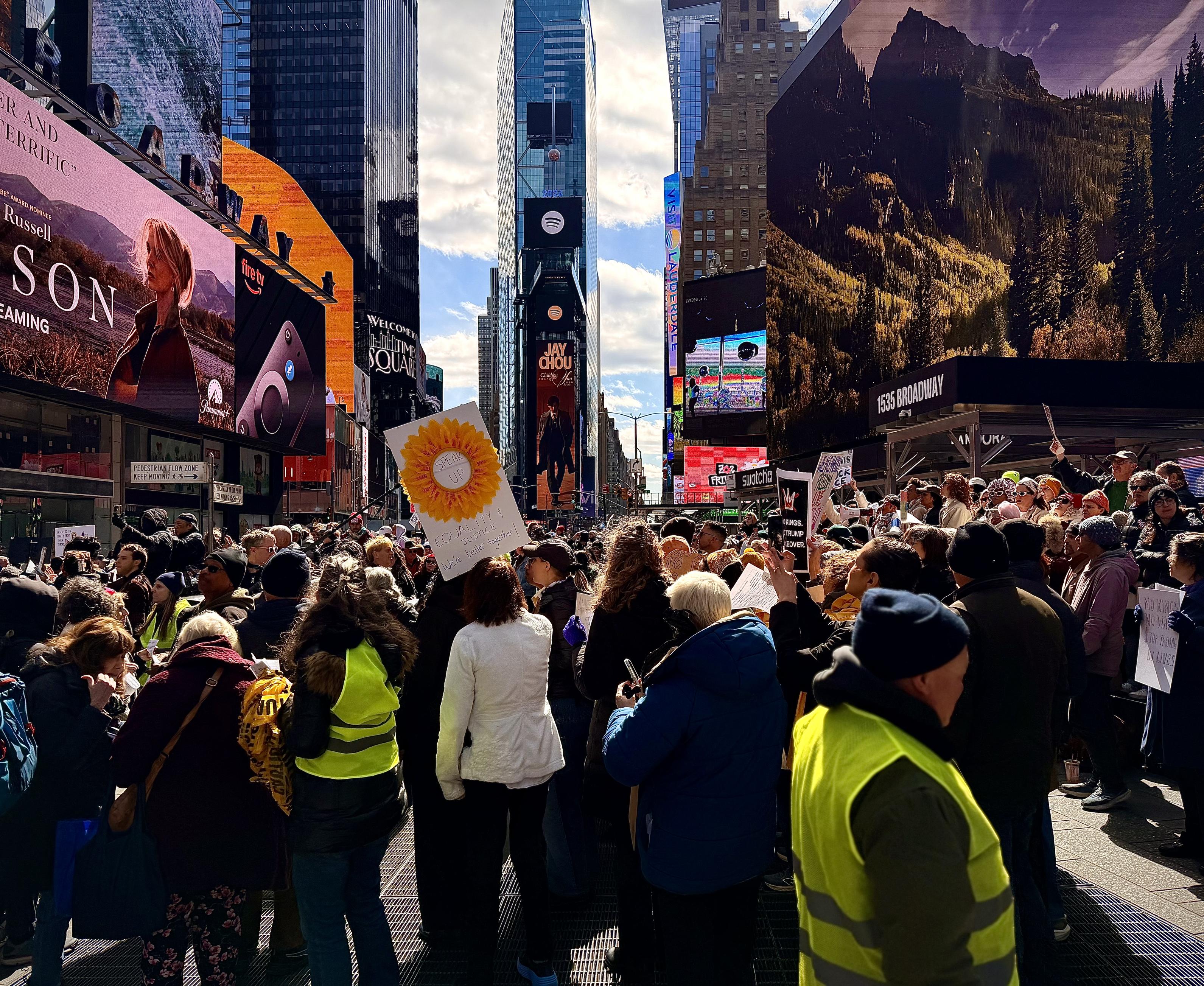 A color, wide-angle photograph that captures a large, dense crowd of protesters gathered in the heart of Times Square, New York City, during the "No Kings" protest on March 28, 2026. The perspective is from behind the crowd, looking north toward the One Times Square building. In the foreground and middle ground, people of diverse ages are wearing heavy winter coats and hats. There are several individuals in the foreground wear bright neon-yellow safety vests as volunteer crowd control. A prominent white hand-drawn sign features a large yellow sunflower and the words "SPEAK UP" in the center, with "EQUALITY & JUSTICE" and "We're better together!" written below. To the right, another person holds a black sign that reads "NO KINGS, TRUMP IS OVER." The bustling atmosphere is framed by massive digital billboards, including an advertisement for Paramount+ on the left and a nature-themed display on the right above the "1535 Broadway" entrance. The urban canyon of Times Square stretches into the background under a bright, partly cloudy sky. Directly in the center of the frame, the iconic One Times Square skyscraper rises above the crowd. At the very summit of this building, the 2026 New Year's Eve Ball is visible as a distinct, glittering geodesic sphere resting atop its flagpole. Just below the ball on the building’s facade, large digital displays show a Spotify logo and a vertical advertisement for Jay Chou.