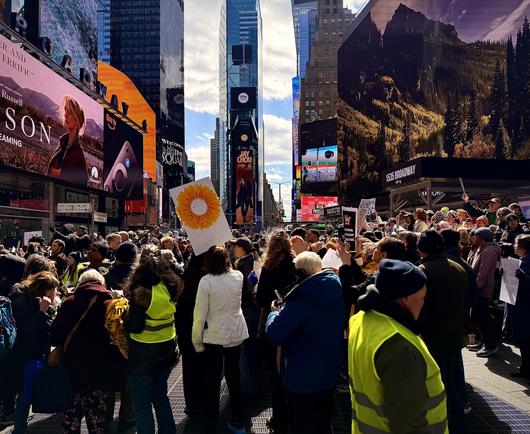 A color, wide-angle photograph that captures a large, dense crowd of protesters gathered in the heart of Times Square, New York City, during the "No Kings" protest on March 28, 2026. The perspective is from behind the crowd, looking north toward the One Times Square building. In the foreground and middle ground, people of diverse ages are wearing heavy winter coats and hats. There are several individuals in the foreground wear bright neon-yellow safety vests as volunteer crowd control. A prominent white hand-drawn sign features a large yellow sunflower and the words "SPEAK UP" in the center, with "EQUALITY & JUSTICE" and "We're better together!" written below. To the right, another person holds a black sign that reads "NO KINGS, TRUMP IS OVER." The bustling atmosphere is framed by massive digital billboards, including an advertisement for Paramount+ on the left and a nature-themed display on the right above the "1535 Broadway" entrance. The urban canyon of Times Square stretches into the background under a bright, partly cloudy sky. Directly in the center of the frame, the iconic One Times Square skyscraper rises above the crowd. At the very summit of this building, the 2026 New Year's Eve Ball is visible as a distinct, glittering geodesic sphere resting atop its flagpole. Just below the ball on the building’s facade, large digital displays show a Spotify logo and a vertical advertisement for Jay Chou.