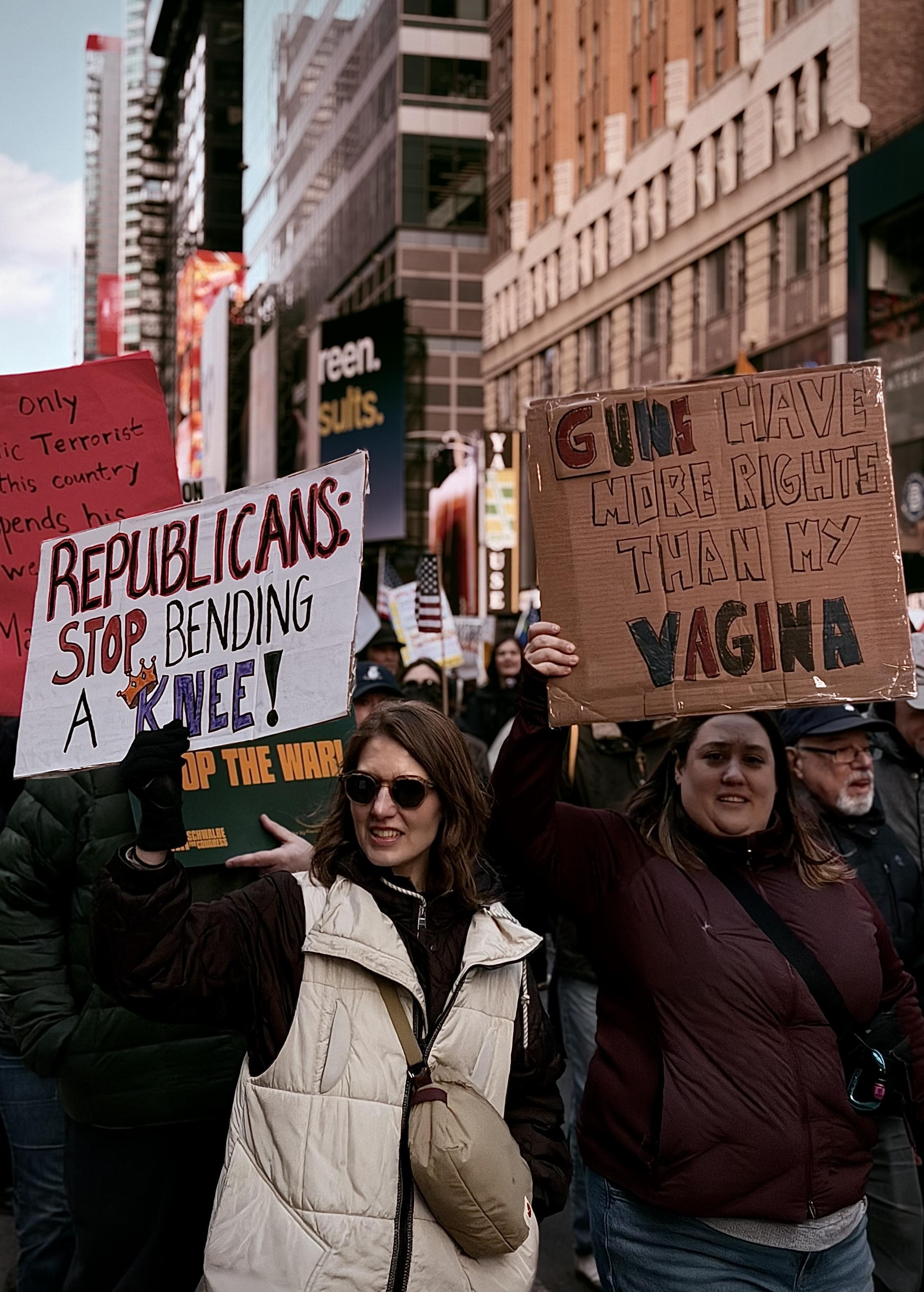 A color, medium-angle photograph in portrait orientation that captures a densely packed No Kings protest march along 7th Avenue in Manhattan. The street is lined with tall, urban buildings and commercial signage. In the foreground, two women lead the way, each holding a hand-drawn cardboard sign. The woman on the left, wearing sunglasses and a white quilted vest over a dark jacket, holds a white sign that reads "REPUBLICANS: STOP BENDING A KNEE!" with a small crown drawn over the word "KNEE." The woman to her right, wearing a maroon puffer jacket, holds a larger cardboard sign that reads "GUNS HAVE MORE RIGHTS THAN MY VAGINA." Their expressions are focused and determined as they walk through the crowd. The background is filled with other protesters, some holding American flags and additional signs, including one that says "STOP THE WAR!" and another red sign partially visible on the left. The lighting is natural and slightly darkened due to nearby buildings blocking afternoon sunlight.