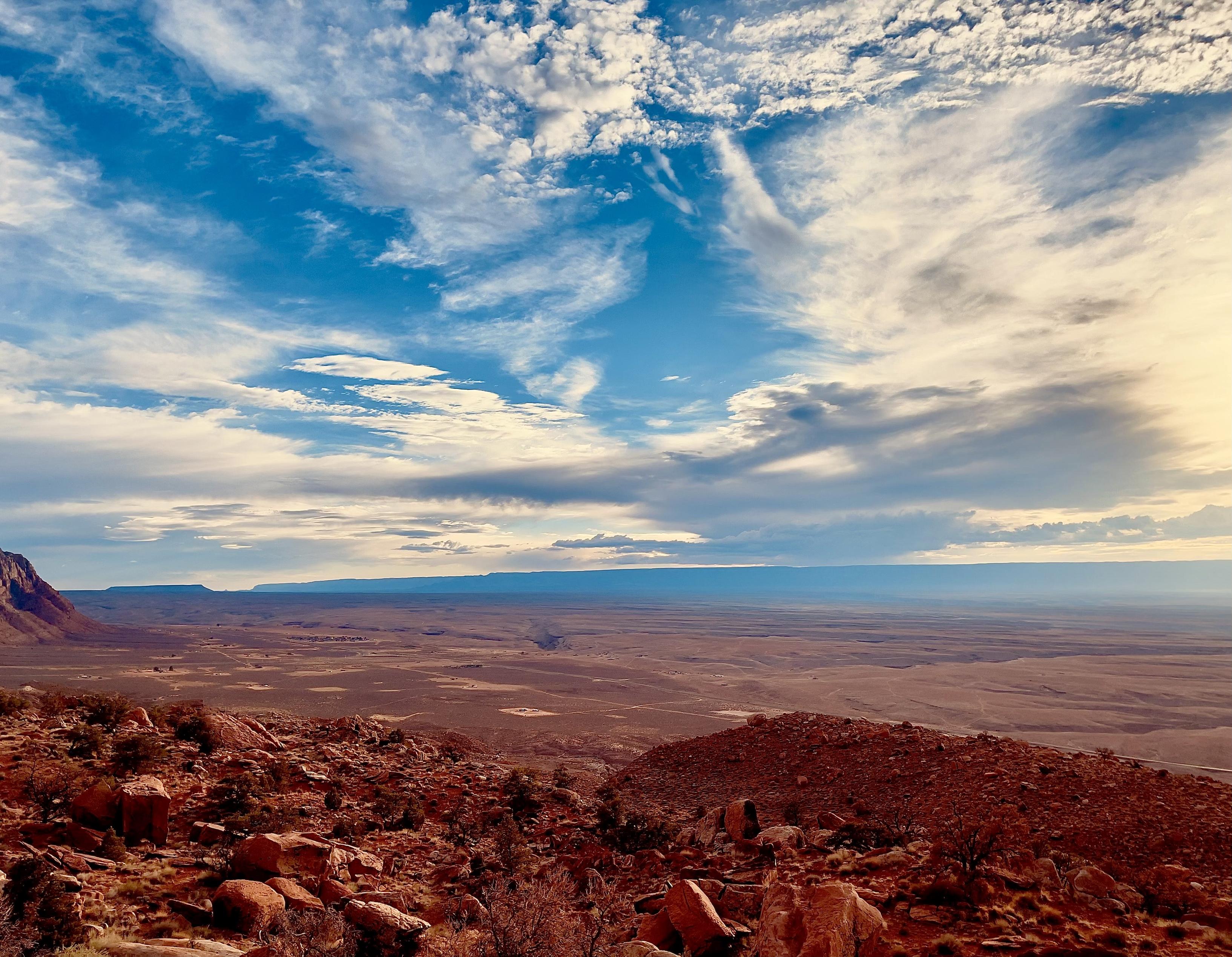 A color, wide-angle photograph in landscape orientation that captures a high-elevation vista overlooking the vast, arid expanse of the Marble Canyon region in northern Arizona under a dramatic sky. In the foreground, a rugged, steep hillside is covered in reddish-brown boulders, fragmented rocks, and sparse, hardy desert vegetation. The earth tones of the foreground are deep and saturated, contrasting sharply with the pale, flat basin that stretches toward the horizon. The middle ground reveals the canyon floor as a sweeping desert plain, marked by subtle geological depressions and distant, isolated structures that appear as tiny specks against the massive landscape. The upper half of the image is dominated by an expansive blue sky filled with a complex tapestry of clouds. Wispy, white cirrus clouds streak across the top, while larger, more shadowed altocumulus formations gather near the horizon, suggesting the approach of a weather front. To the right, the sun’s glow permeates the clouds, casting a soft, hazy light over the distant, flat-topped mesas and plateaus that define the horizon line. 