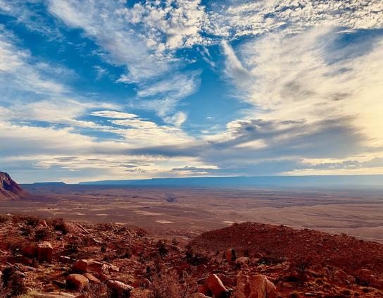 A color, wide-angle photograph in landscape orientation that captures a high-elevation vista overlooking the vast, arid expanse of the Marble Canyon region in northern Arizona under a dramatic sky. In the foreground, a rugged, steep hillside is covered in reddish-brown boulders, fragmented rocks, and sparse, hardy desert vegetation. The earth tones of the foreground are deep and saturated, contrasting sharply with the pale, flat basin that stretches toward the horizon. The middle ground reveals the canyon floor as a sweeping desert plain, marked by subtle geological depressions and distant, isolated structures that appear as tiny specks against the massive landscape. The upper half of the image is dominated by an expansive blue sky filled with a complex tapestry of clouds. Wispy, white cirrus clouds streak across the top, while larger, more shadowed altocumulus formations gather near the horizon, suggesting the approach of a weather front. To the right, the sun’s glow permeates the clouds, casting a soft, hazy light over the distant, flat-topped mesas and plateaus that define the horizon line. 