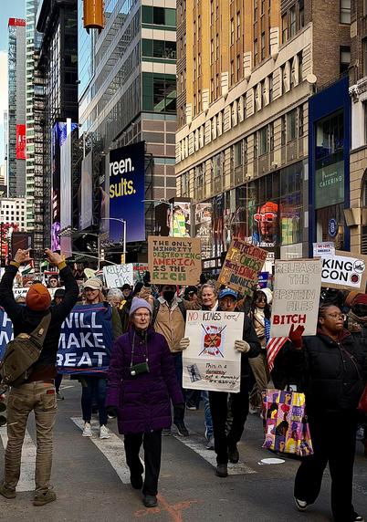 A color, wide-angle photograph in portrait orientation that captures the No Kings protest march on Seventh Avenue near 40th Street in the Garment District neighborhood of Manhattan on March 28, 2026. In the foreground, a diverse group of demonstrators marches toward the camera. A woman in a vibrant purple puffer jacket and lavender beanie walks centered in the frame, flanked by others holding handmade signs. One prominent sign features a red "X" over a King of Diamonds playing card with the text "NO KINGS here! This is a democracy not a monarchy or a dictatorship!!!" To her right, a woman in a black jacket and red gloves holds a sign that reads "RELEASE THE EPSTEIN FILES BITCH" in bold red and black lettering. Other visible signs include messages like "THE ONLY MINORITY DESTROYING AMERICA IS THE RICH" and "RELEASE THE FILES." The background showcases the urban canyon of Midtown Manhattan just south of Times Square, with towering glass and stone skyscrapers lining the street under a clear sky. On the right, the facade of a building features a "Paris Baguette Catering" sign and a large advertisement with a person wearing bright red-rimmed glasses. To the far left, a man with a tan backpack and orange beanie holds up a smartphone to record the crowd. Late afternoon sunlight is filtering onto the scene.