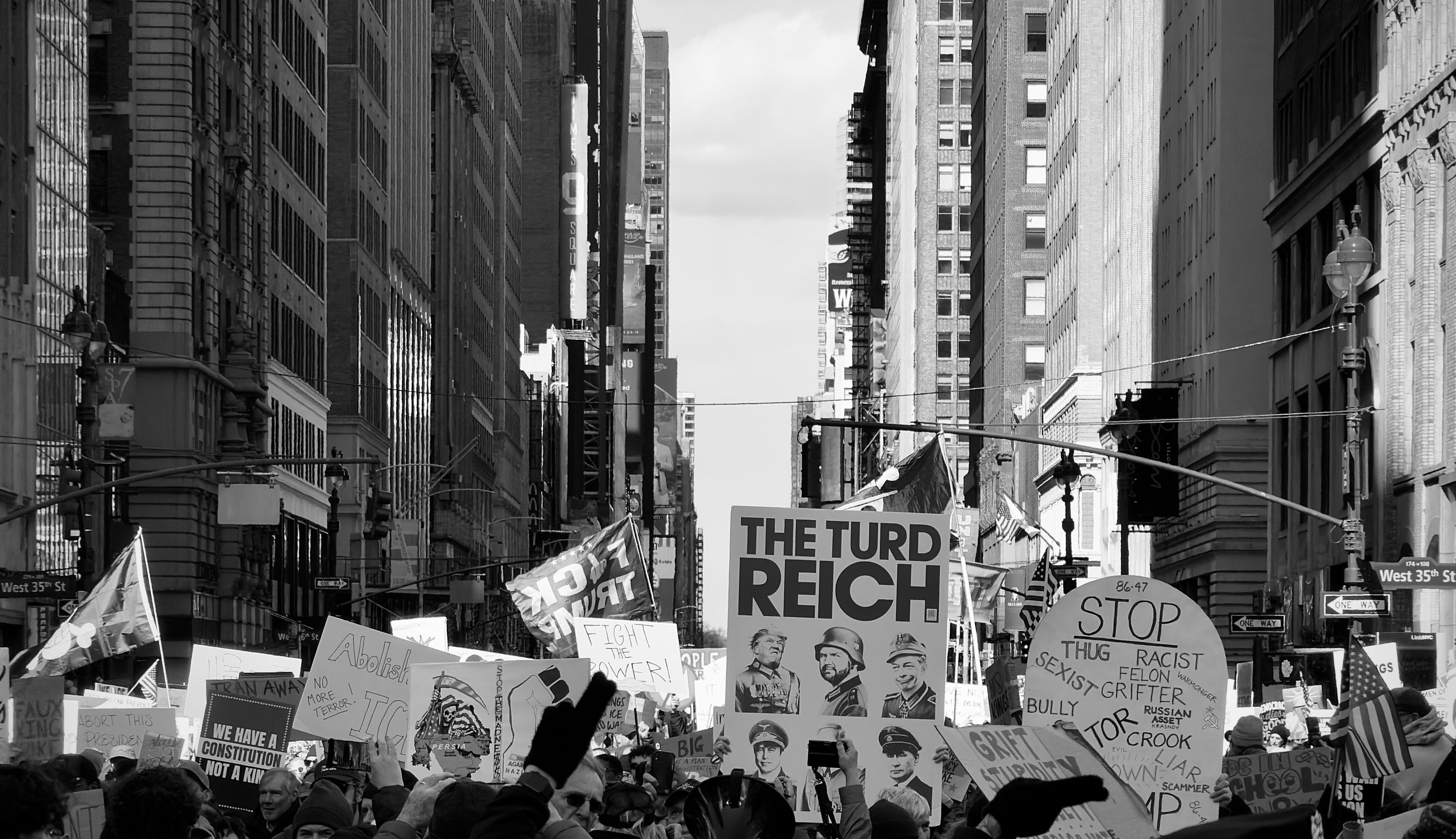 A noir, wide-angle photograph in landscape orientation that captures a dense crowd of demonstrators during the No Kings protest on March 28, 2026, looking north from 7th Avenue at the intersection of West 35th Street in Manhattan. Tall skyscrapers line both sides of the street, creating a deep urban canyon under a bright, mostly clear sky. In the foreground and middle ground, numerous protesters hold a variety of signs and flags. A prominent rectangular poster in the center reads "THE TURD REICH" in bold letters, featuring six caricatured portraits of political figures in military-style uniforms. Nearby, a large circular sign lists a series of grievances, including terms like "THUG," "RACIST," "SEXIST," and "GRIFTER." The atmosphere of the rally is captured through a mix of handmade and printed materials reflecting a range of social and political concerns. The crowd is shown from the chest up, with many individuals holding their signs high, their backs to the camera as they face toward the heart of Midtown. Late afternoon sun is casting long shadows across the scene.
