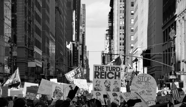 A noir, wide-angle photograph in landscape orientation that captures a dense crowd of demonstrators during the No Kings protest on March 28, 2026, looking north from 7th Avenue at the intersection of West 35th Street in Manhattan. Tall skyscrapers line both sides of the street, creating a deep urban canyon under a bright, mostly clear sky. In the foreground and middle ground, numerous protesters hold a variety of signs and flags. A prominent rectangular poster in the center reads "THE TURD REICH" in bold letters, featuring six caricatured portraits of political figures in military-style uniforms. Nearby, a large circular sign lists a series of grievances, including terms like "THUG," "RACIST," "SEXIST," and "GRIFTER." The atmosphere of the rally is captured through a mix of handmade and printed materials reflecting a range of social and political concerns. The crowd is shown from the chest up, with many individuals holding their signs high, their backs to the camera as they face toward the heart of Midtown. Late afternoon sun is casting long shadows across the scene.