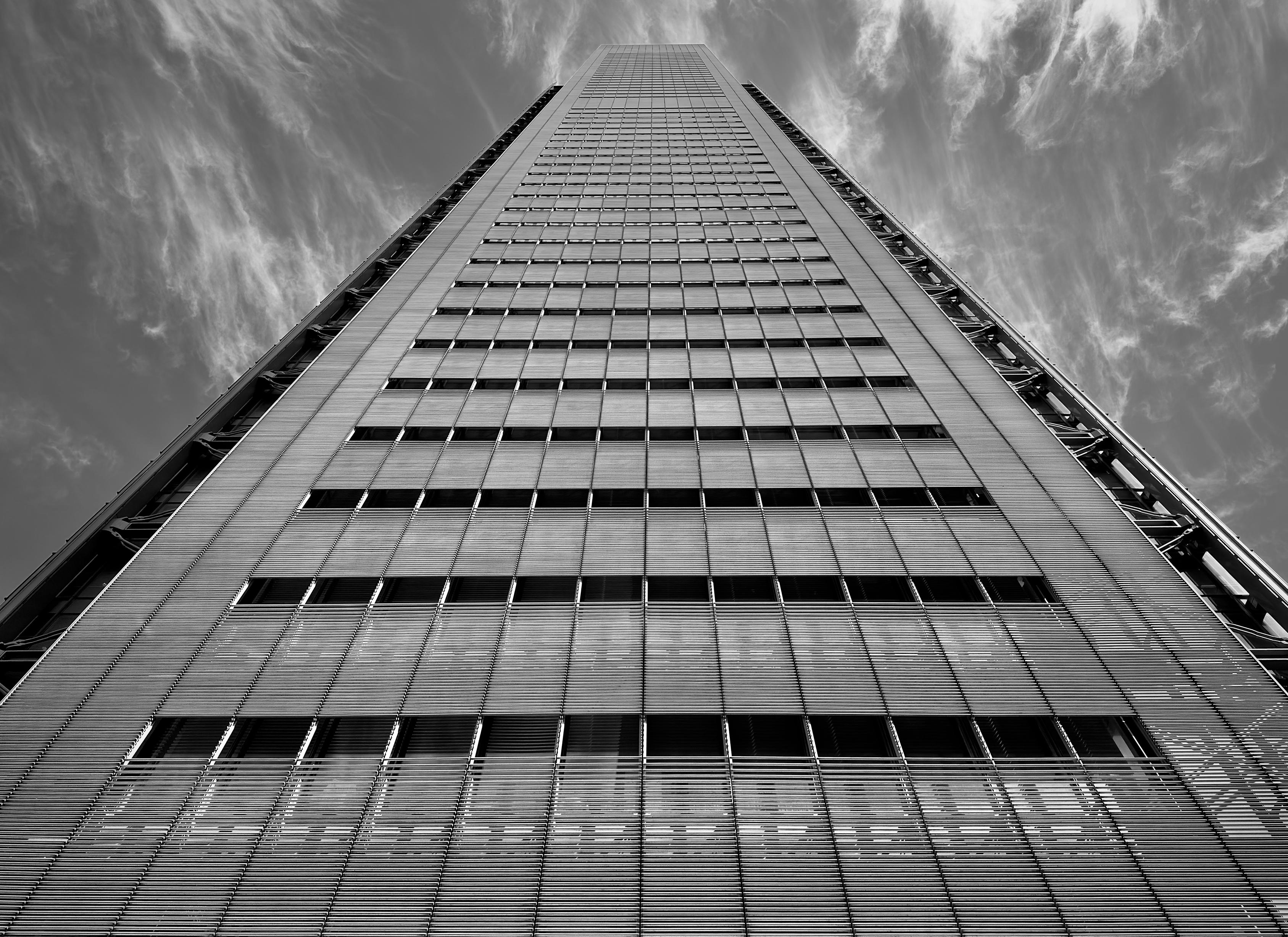 A monochrome, low-angle photograph that captures the south facade of the New York Times Building in the Garment District neighborhood of Manhattan, looking directly upward toward the sky. The perspective creates a powerful sense of verticality, as the building’s parallel lines converge toward a central point high above, making the skyscraper appear to pierce the wispy, streaked clouds. The facade is defined by a rhythmic, grid-like pattern of glass windows layered behind a distinctive curtain of horizontal ceramic rods, which create a fine, textured screen across the building's surface. The high-contrast black-and-white tones emphasize the play of light and shadow across the architectural details. Exposed steel framing is visible along the outer edges of the tower, highlighting its industrial aesthetic and structural transparency. The sky is a deep, dramatic grey, filled with feathery cirrus clouds that radiate outward from behind the building’s crown, adding a dynamic, ethereal quality to the rigid geometric precision of the skyscraper.
