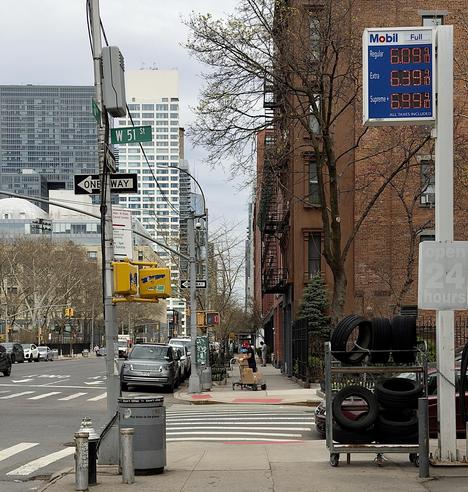 A color, medium-angle photograph in square aspect ratio that captures the street scene along 51st Street in the Hell’s Kitchen neighborhood of Manhattan, focused on a Mobil gas station at an intersection. In the foreground, a prominent white sign displays fuel prices in bright red and blue LED digits: $6.09 for Regular, $6.39 for Extra, and $6.99 for Supreme. To the right of the price sign, a tiered metal rack holds several black tires, and a weathered, vertical "open 24 hours" sign is visible. The mid-ground features a gray Range Rover parked along the curb and a delivery person in a red cap pushing a hand truck laden with cardboard boxes down the sidewalk. Street signs for W 51 St and a "One Way" indicator are mounted on a utility pole alongside a yellow traffic signal. The background is a mix of classic New York architecture, including a multi-story brownstone with black fire escapes and modern, white high-rise buildings that tower over the city blocks under a party cloudy, overcast sky.