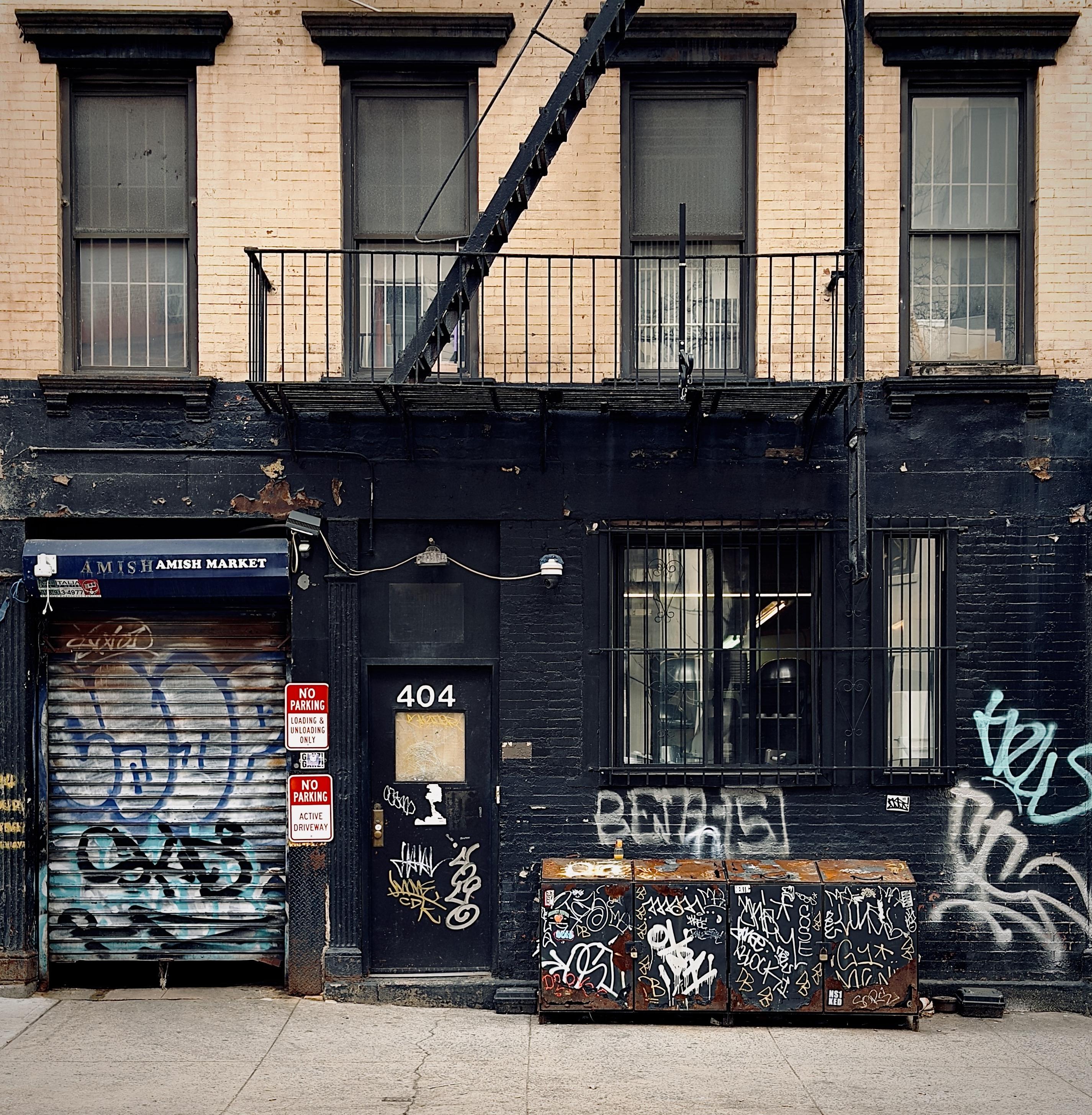 A color, medium-angle photograph in square aspect ratio that captures the street scene along 50th Street in the Hell’s Kitchen neighborhood of Manhattan. The focal point is a building facade that features a striking contrast between its tan brick upper floor with four windows, and a matte black painted ground floor. A black metal fire escape ladder descends diagonally across the upper windows toward a second-story balcony. On the ground level, the storefront for "Amish Market" is secured by a rusted, silver rolling metal gate heavily covered in blue and black graffiti. To the right of the gate, a black door is labeled with the number 404 and is adorned with various white and yellow street art tags. Two red and white "No Parking" signs are mounted on the wall between the gate and the door. The right side of the ground floor contains a large, barred window through which interior shop lights and equipment are visible. Below this window sits a row of four interconnected, rusted metal refuse storage bins, each completely covered in intricate white and black graffiti tags. The surrounding black brick walls are further marked by large, light-colored spray-painted tags and peeling paint. The sidewalk in the foreground is a light gray concrete, showing typical wear and minor cracks, completing the gritty, industrial aesthetic of the city scene. An overcast afternoon sky is evenly illuminating the scene.