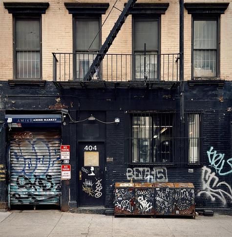 A color, medium-angle photograph in square aspect ratio that captures the street scene along 50th Street in the Hell’s Kitchen neighborhood of Manhattan. The focal point is a building facade that features a striking contrast between its tan brick upper floor with four windows, and a matte black painted ground floor. A black metal fire escape ladder descends diagonally across the upper windows toward a second-story balcony. On the ground level, the storefront for "Amish Market" is secured by a rusted, silver rolling metal gate heavily covered in blue and black graffiti. To the right of the gate, a black door is labeled with the number 404 and is adorned with various white and yellow street art tags. Two red and white "No Parking" signs are mounted on the wall between the gate and the door. The right side of the ground floor contains a large, barred window through which interior shop lights and equipment are visible. Below this window sits a row of four interconnected, rusted metal refuse storage bins, each completely covered in intricate white and black graffiti tags. The surrounding black brick walls are further marked by large, light-colored spray-painted tags and peeling paint. The sidewalk in the foreground is a light gray concrete, showing typical wear and minor cracks, completing the gritty, industrial aesthetic of the city scene. An overcast afternoon sky is evenly illuminating the scene.