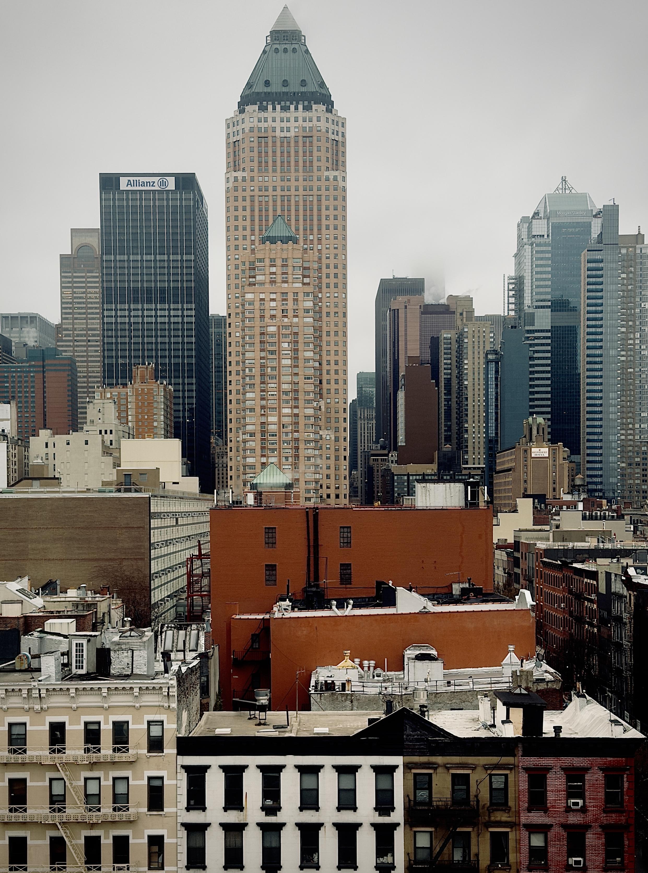 A color, wide-angle photograph in portrait orientation that captures a high-angle, eastward-facing view from Tenth Avenue, looking across the rooftops of Hell’s Kitchen toward the dense skyscrapers of the Theater District in midtown Manhattan. In the foreground, a row of traditional multi-story tenement buildings displays a mix of architectural textures, including beige masonry, white facades with black-trimmed windows, and red brick. Their rooftops are cluttered with quintessential New York details: silver vents, water tanks, black chimneys, and industrial HVAC units. Centrally, a large, windowless orange-brick side wall of a mid-rise building provides a bold block of color that anchors the lower half of the frame against the gray, overcast afternoon sky. Rising sharply behind the low-rise foreground is a crowded skyline of iconic commercial towers. The most prominent is the One Worldwide Plaza, a tan, postmodern skyscraper easily identified by its distinct pyramidal roof topped with a glass lantern. To its left stands the dark, grid-like facade of the Allianz building, and to the right, the sleek, glass-clad Morgan Stanley building with its tiered, metallic crown. Other vertical structures in shades of navy, gray, and brown fill the gaps, creating a tightly packed wall of steel and glass. A light mist partially obscures some of the taller mid-block buildings, adding a moody, atmospheric depth to the urban landscape.