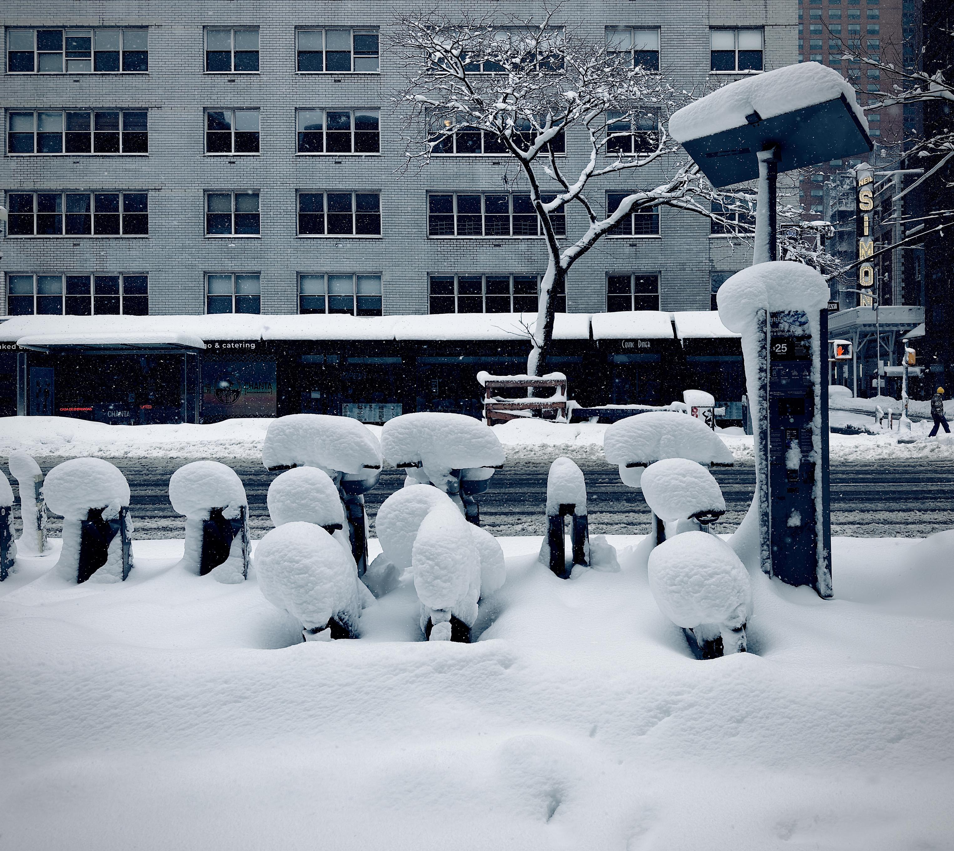 A color, wide-angle photograph taken along Eighth Avenue in the Hell’s Kitchen neighborhood of Manhattan that captures a heavy winter snowstorm. In the foreground, a row of Citi Bikes is nearly consumed by thick, pillowy drifts of white snow. The bicycles are parked in their docking stations, each capped with a rounded, mushroom-like mound of snow several inches deep. To the right, a tall, rectangular bike-share kiosk stands with a flat, solar-panel roof also heavily weighed down by a thick layer of snow. The ground is a seamless, undisturbed blanket of white, leading back toward the dark, slushy asphalt of the street. In the background, a large, multi-story brick apartment building with rows of dark, grid-patterned windows dominates the view. At the street level, storefronts with dark awnings are visible, including a sign for the "Cosmic Diner." To the right, the vertical neon "SIMON" sign for the Neil Simon Theatre glows faintly against the gray, overcast sky. A bare, snow-dusted tree reaches upward in front of the building, its dark branches providing a stark contrast to the white architecture. The overall atmosphere is quiet and cold, with a muted, cool-toned color palette dominated by whites, grays, and deep blues.