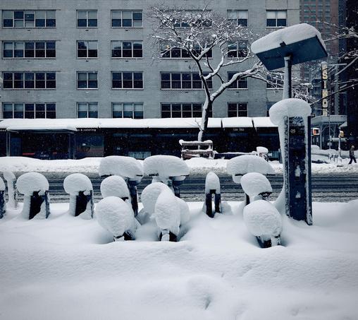 A color, wide-angle photograph taken along Eighth Avenue in the Hell’s Kitchen neighborhood of Manhattan that captures a heavy winter snowstorm. In the foreground, a row of Citi Bikes is nearly consumed by thick, pillowy drifts of white snow. The bicycles are parked in their docking stations, each capped with a rounded, mushroom-like mound of snow several inches deep. To the right, a tall, rectangular bike-share kiosk stands with a flat, solar-panel roof also heavily weighed down by a thick layer of snow. The ground is a seamless, undisturbed blanket of white, leading back toward the dark, slushy asphalt of the street. In the background, a large, multi-story brick apartment building with rows of dark, grid-patterned windows dominates the view. At the street level, storefronts with dark awnings are visible, including a sign for the "Cosmic Diner." To the right, the vertical neon "SIMON" sign for the Neil Simon Theatre glows faintly against the gray, overcast sky. A bare, snow-dusted tree reaches upward in front of the building, its dark branches providing a stark contrast to the white architecture. The overall atmosphere is quiet and cold, with a muted, cool-toned color palette dominated by whites, grays, and deep blues.